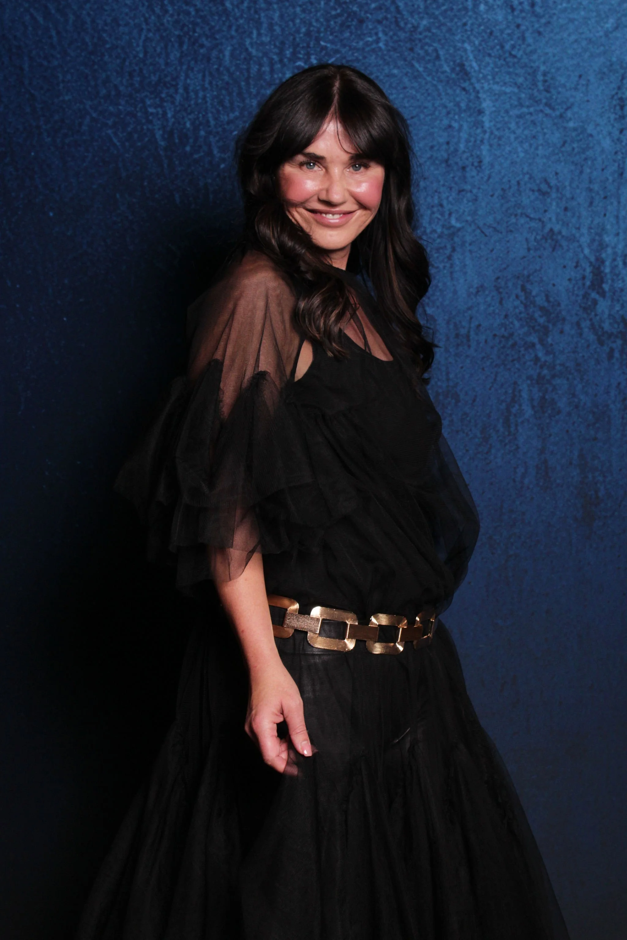 A woman wearing a black dress with sheer, ruffled sleeves, a gold chain belt, and smiling against a dark blue textured background.