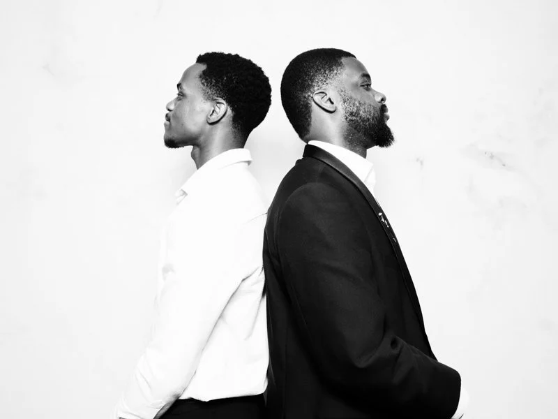 Two men standing back to back, facing opposite directions, against a plain white background.