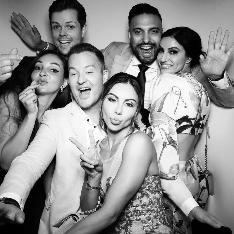 Group of six friends smiling, making playful gestures, and posing for a photo together, dressed in stylish clothing, in black and white.