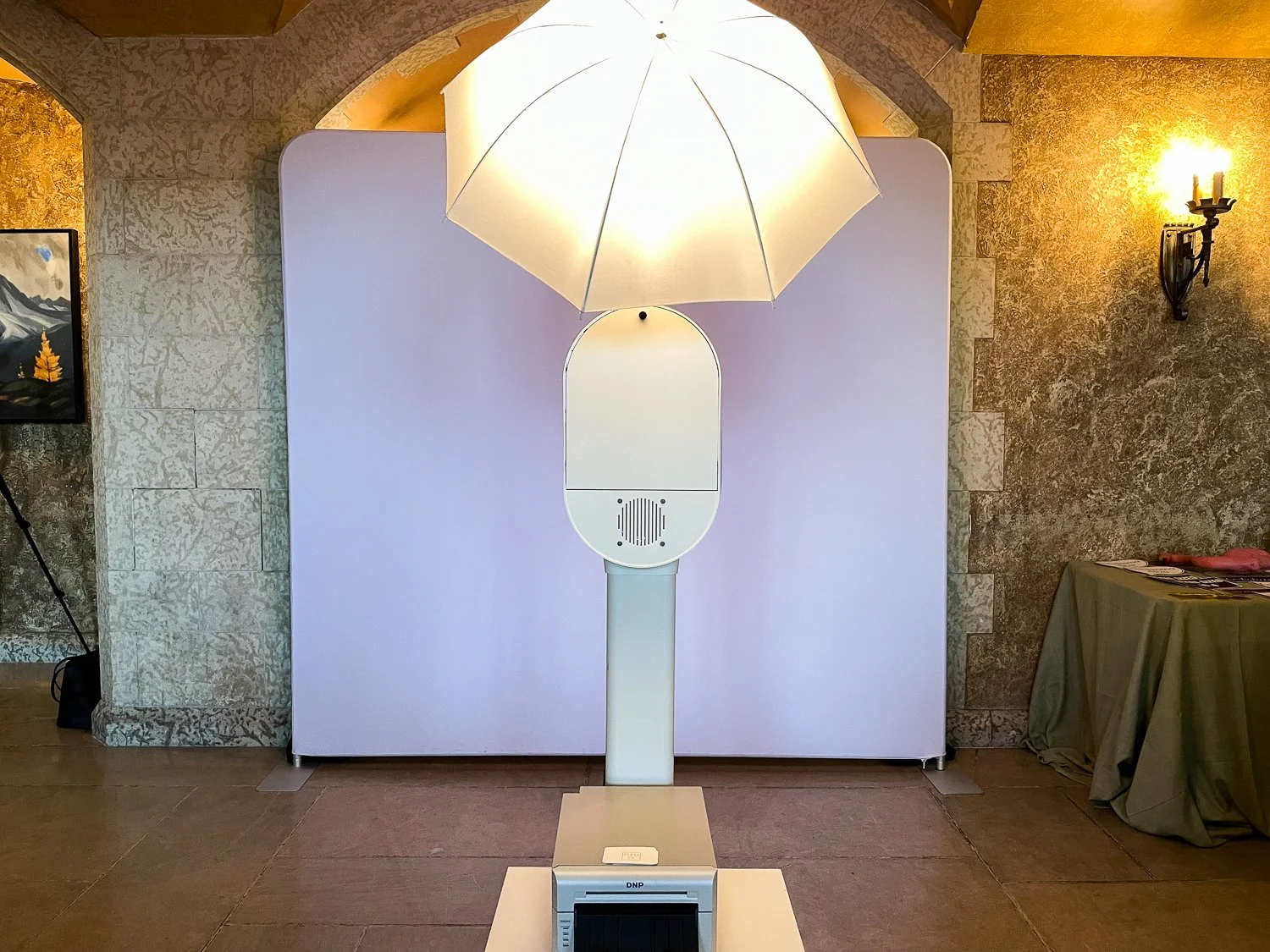 A white balloon-shaped device with a circular mirror and speaker grill stands in front of a plain white background, illuminated by a white umbrella light, in an indoor setting with stone walls and a table covered with a beige cloth to the right.