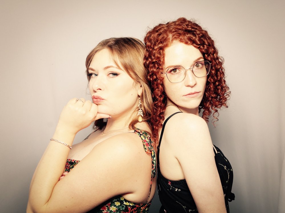 Two women with shoulder-length hair, one with curly red hair and glasses, and the other with straight light brown hair, posing back-to-back against a plain background