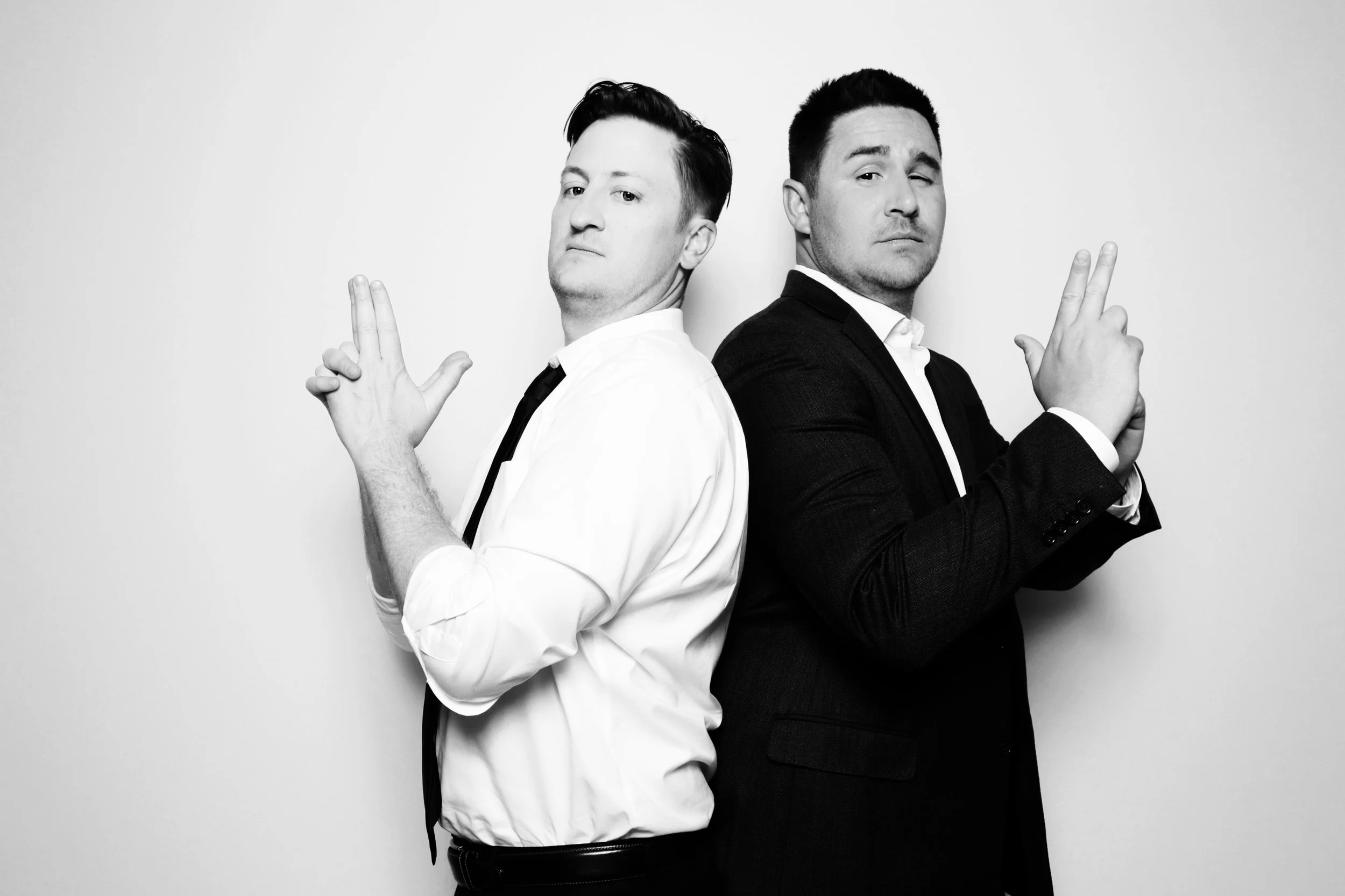 Two men standing back-to-back, facing sideways, making peace signs with their hands, dressed in suits, in black and white photo.