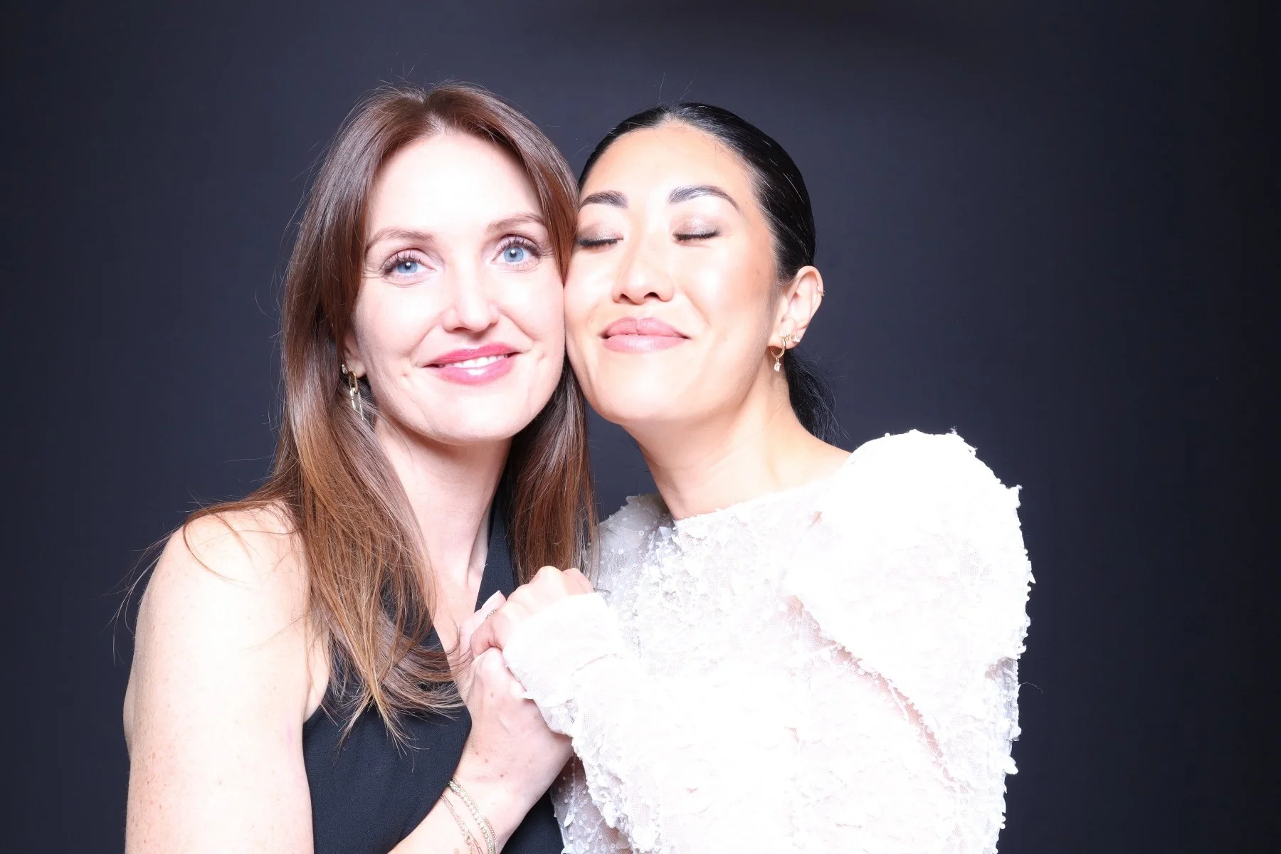 Two women smiling and holding hands against a dark background, one with brown hair and blue eyes, and the other with black hair and closed eyes.