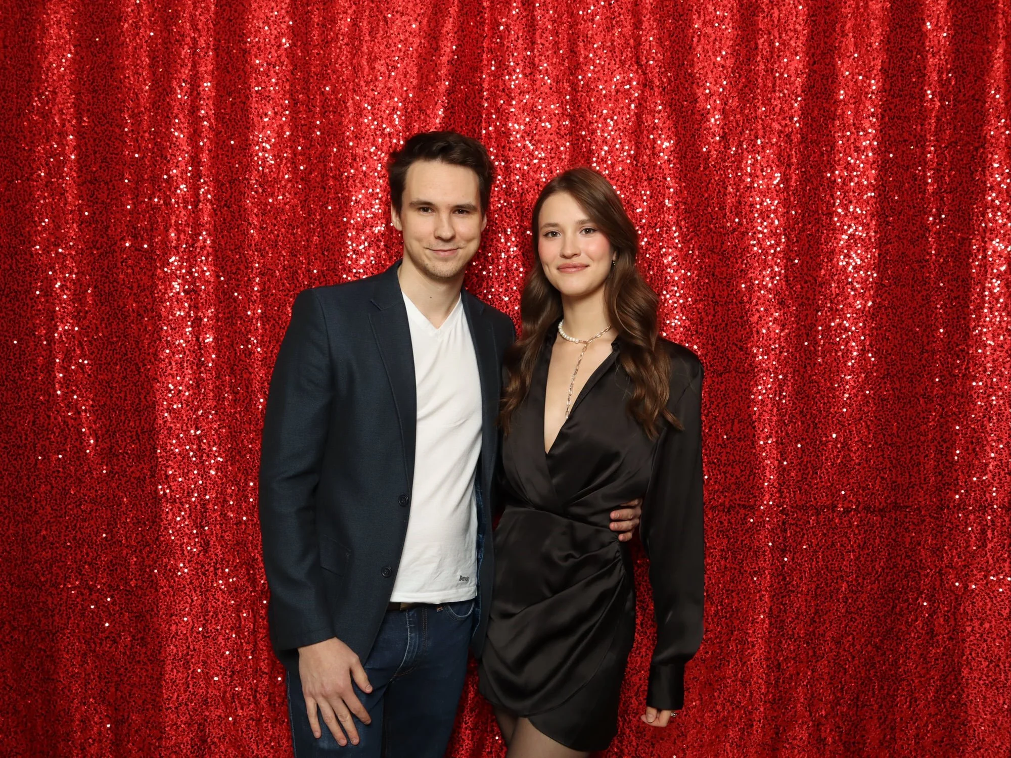 A young man and woman standing close together in front of a red, glittery backdrop, smiling at the camera. The man is wearing a dark blazer over a white t-shirt, and the woman is dressed in a black satin blouse with a pearl necklace.