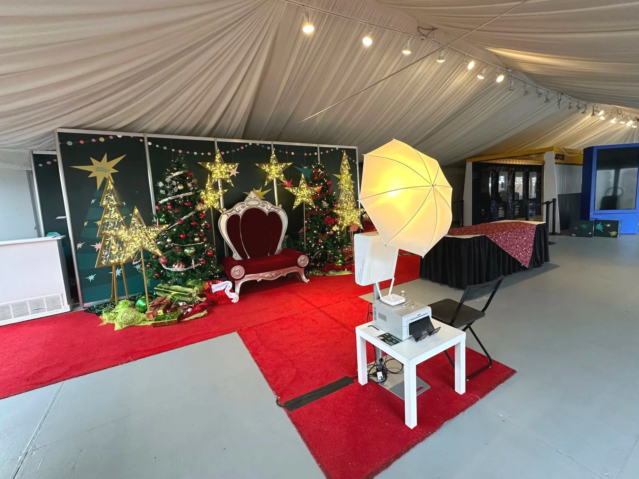Christmas stage setup with decorated Christmas trees, gold star decorations, and a red velvet throne. A photographer's lighting and equipment are set up in the foreground. The scene is inside a large tent with a draped ceiling and string lights.