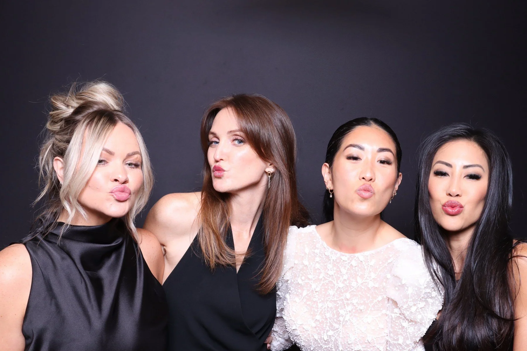Four women with different hair styles and colors, pressing their lips together in a playful pose against a dark background.