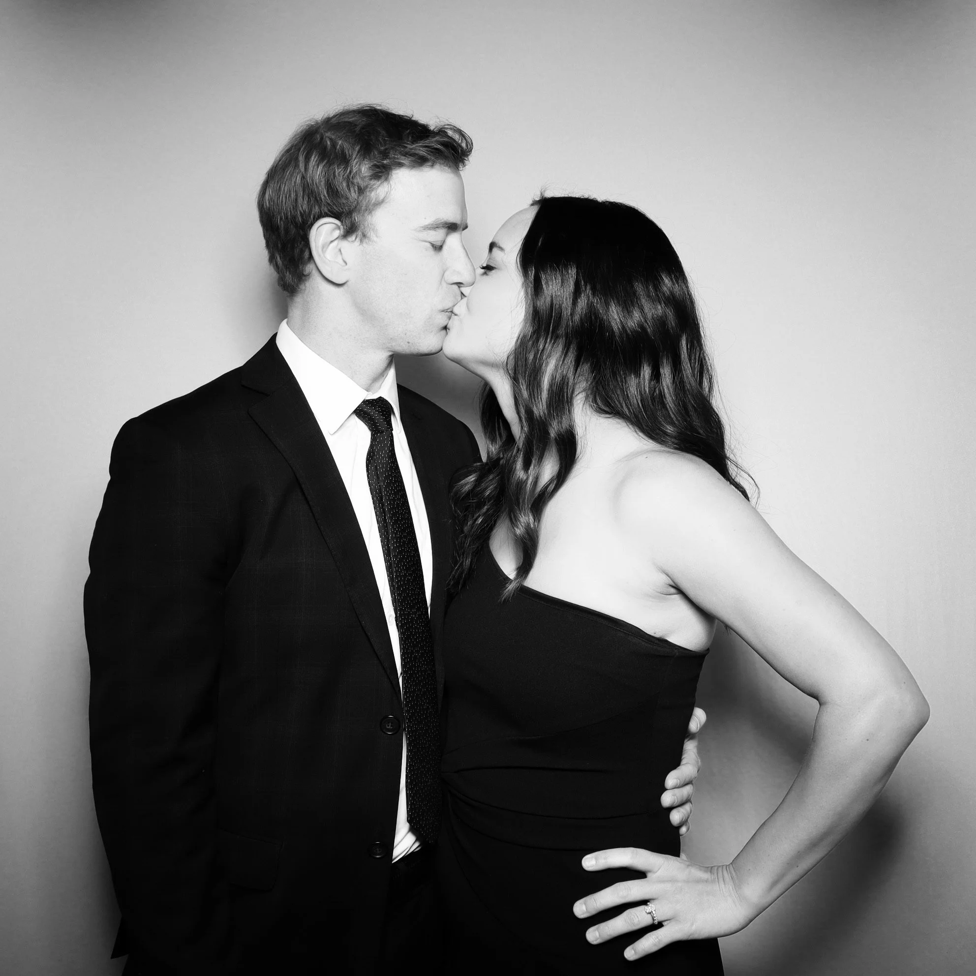 A black and white photo of a couple kissing, with the man in a suit and tie, and the woman in a strapless dress.