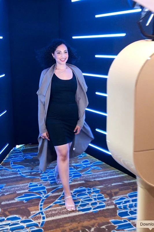 A woman with curly black hair smiling in a modern room with blue neon lighting, wearing a black dress, gray coat, and high heels.