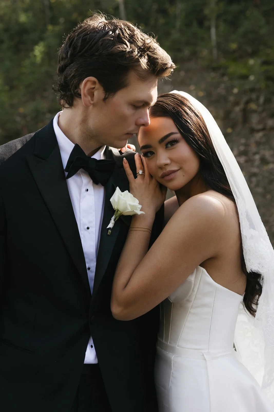 A bride and groom embrace outdoors during their wedding, with the groom in a black tuxedo and the bride in a white wedding dress and veil.
