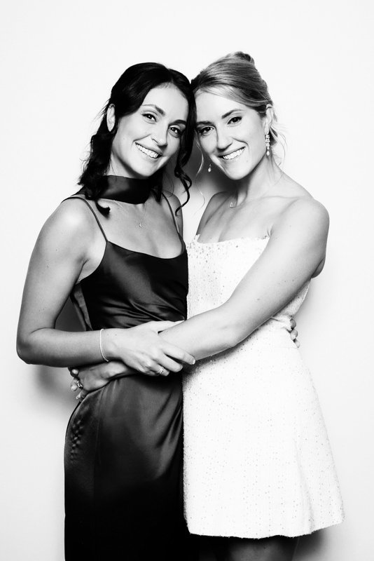 Two women in formal dresses standing closely together, smiling at the camera against a plain background.