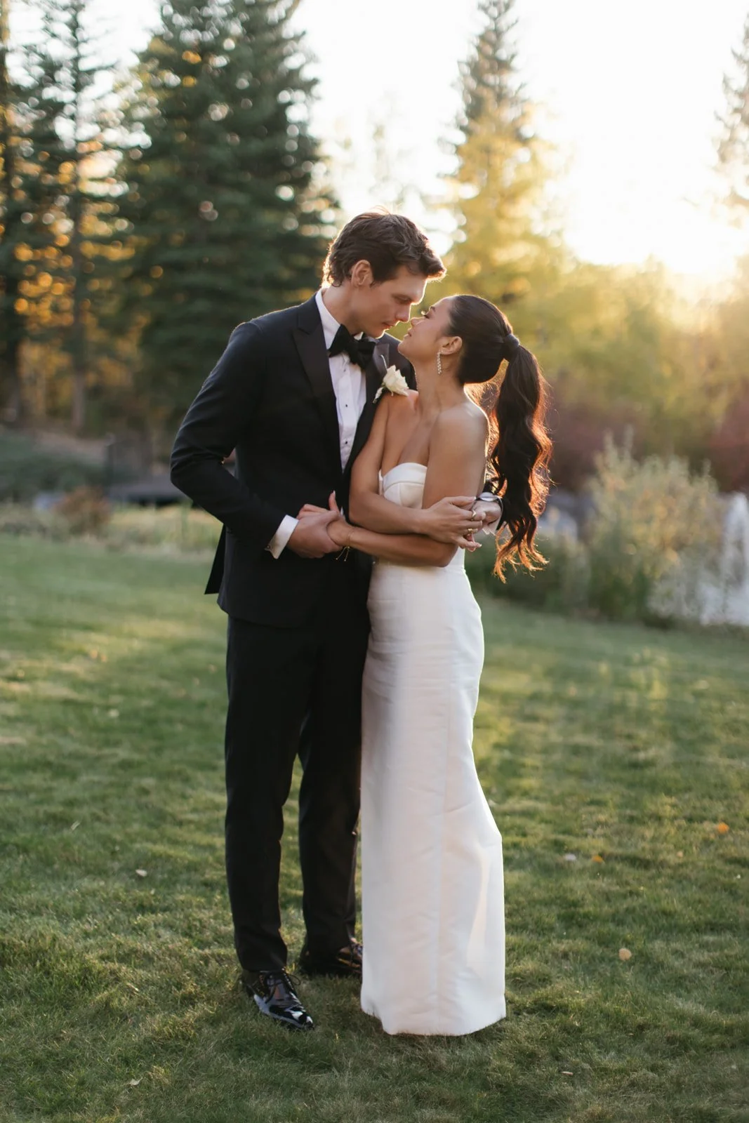 A bride and groom stand close together outdoors during sunset. The groom wears a black tuxedo, and the bride wears a strapless white wedding gown. They are gazing into each other's eyes, with the bride holding the groom's arm.