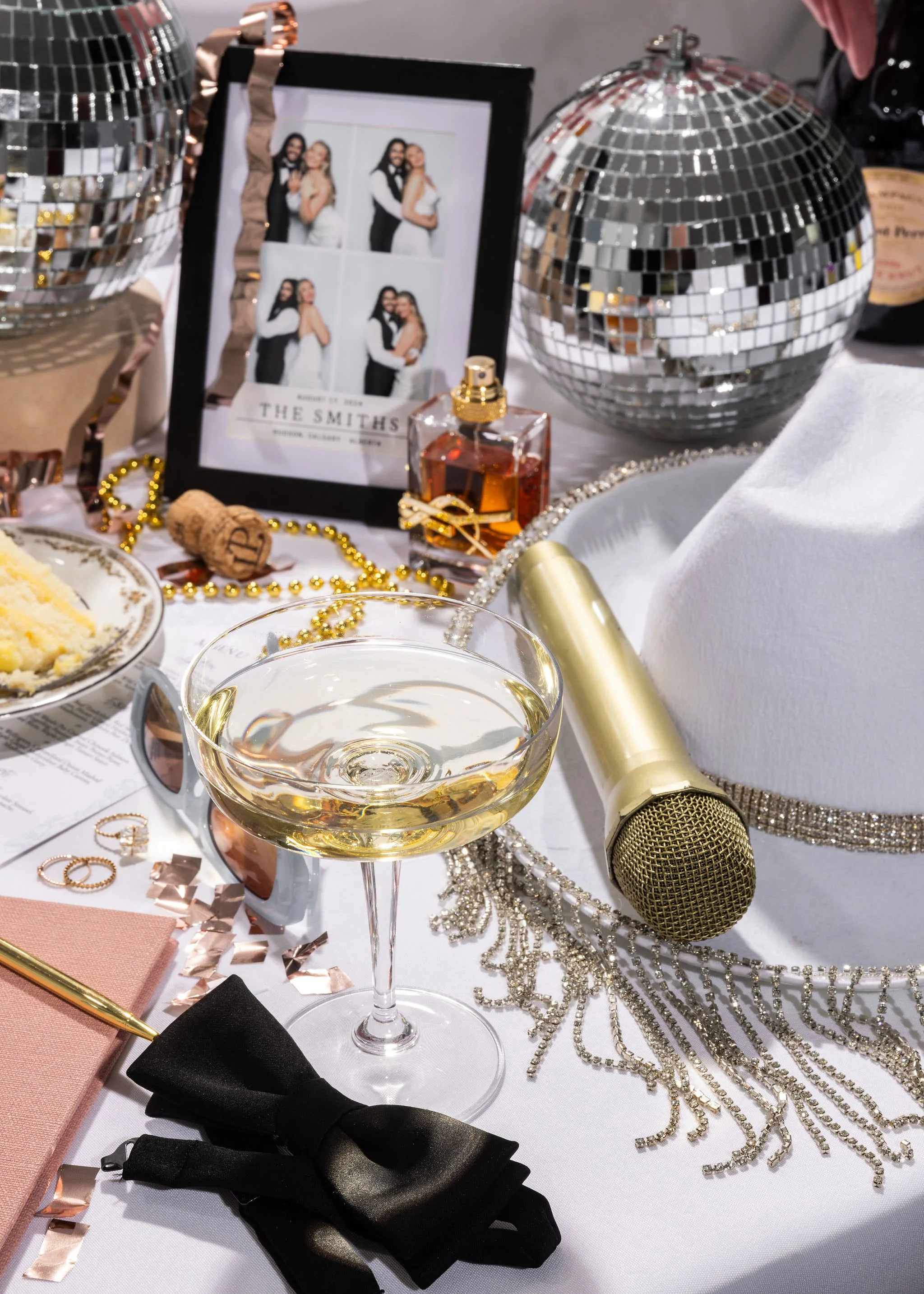 A festive table setting with a microphone, a glass of white wine, champagne glasses, disco balls, a photo frame with images of three women, a white hat with a rhinestone band, a gold pen, a black bow tie, jewelry, a small bottle of perfume, and a pla