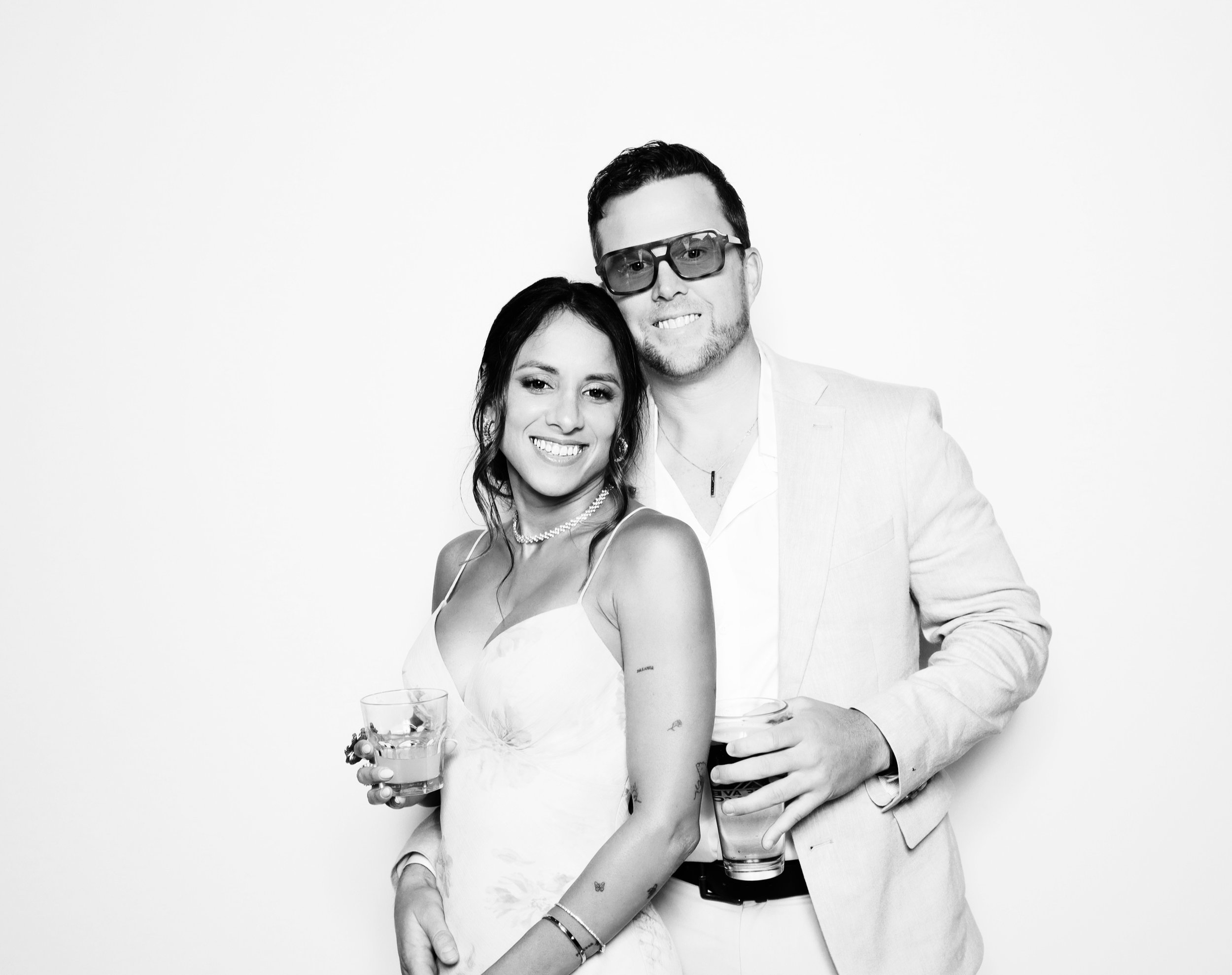 A smiling woman and man, both dressed in light-colored clothes, holding drinks and standing close together against a plain background.