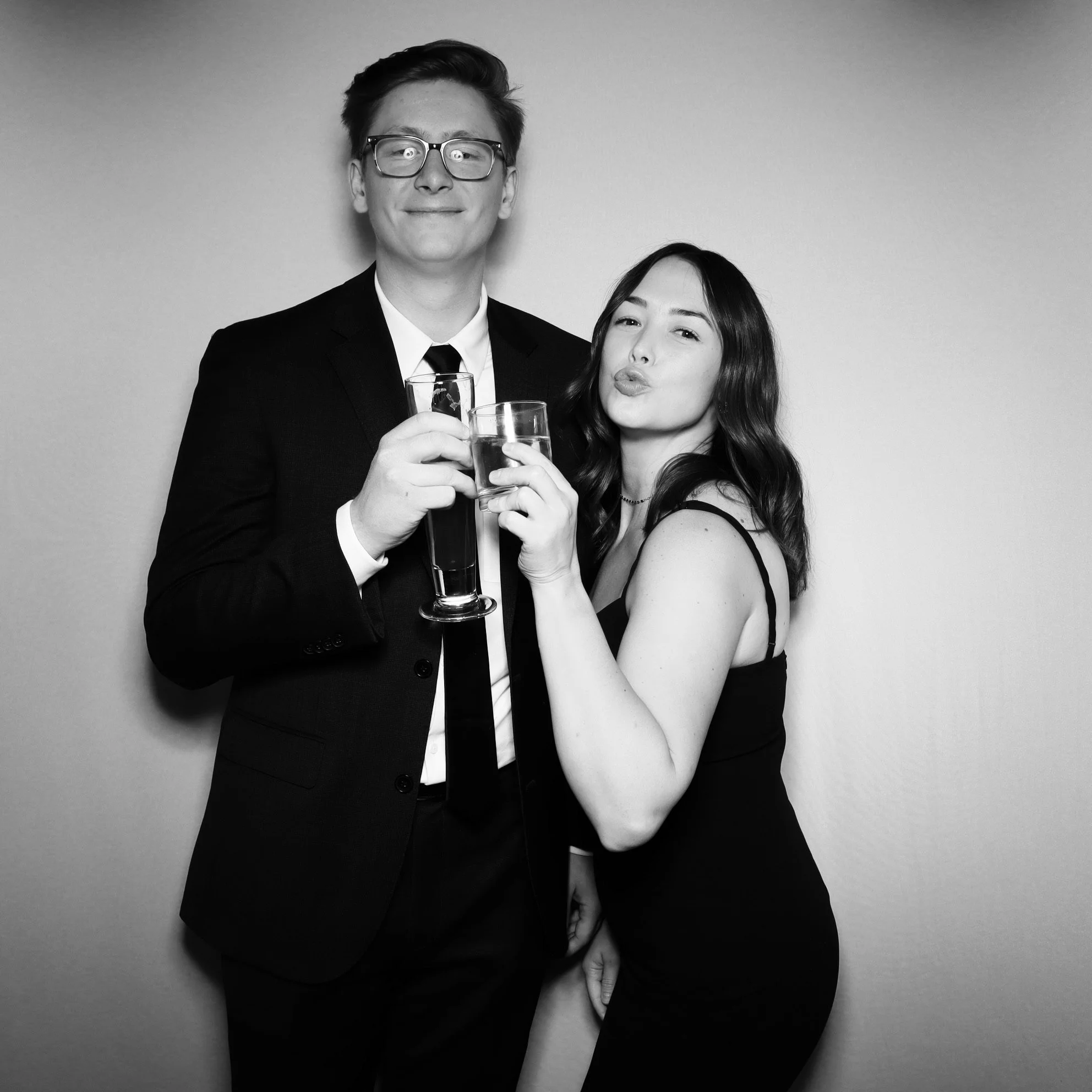 Black and white photo of a young man and woman at a celebration, holding drinks and posing for the camera.