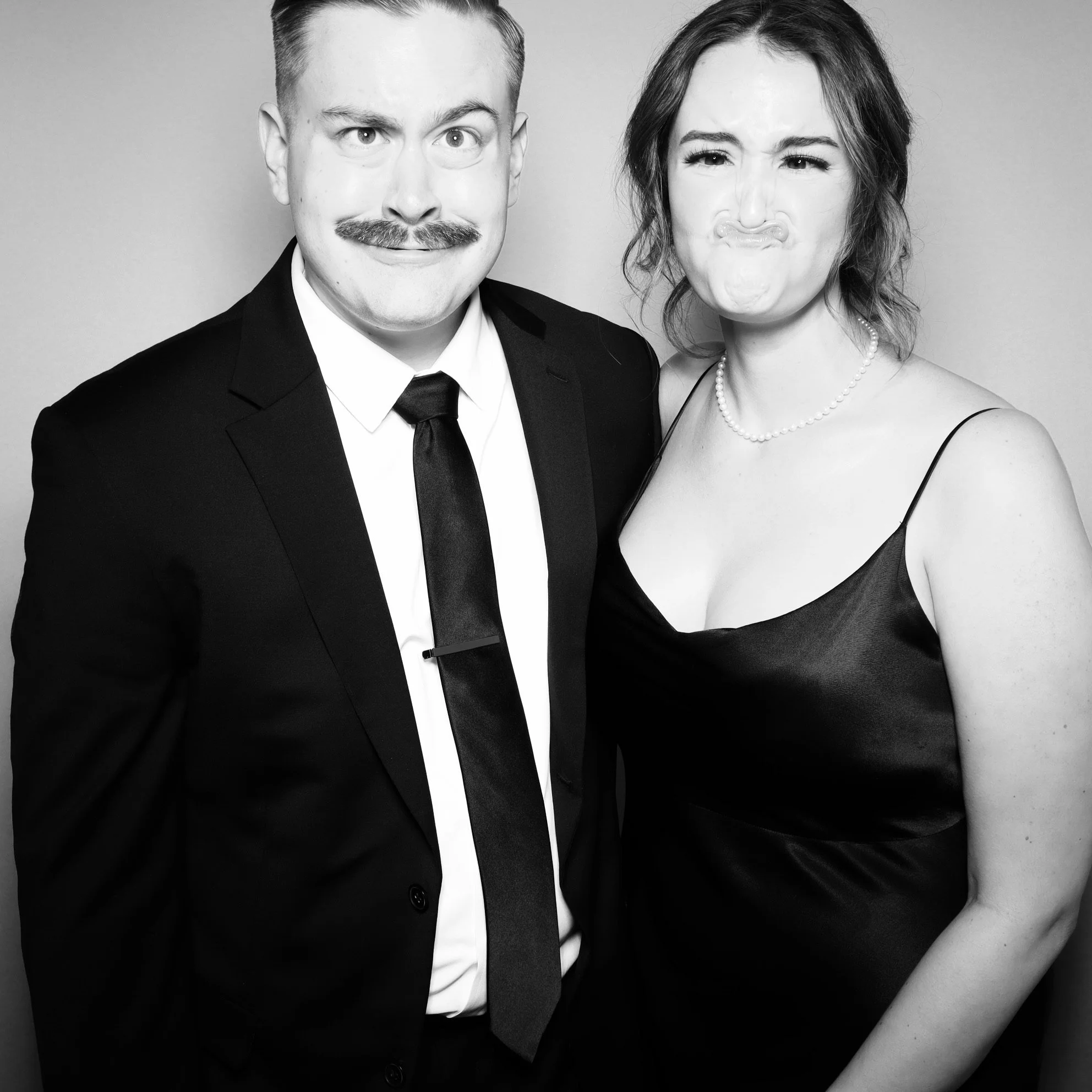 A black-and-white photo of a man and woman making silly faces at the camera. The man is wearing a suit and tie with a mustache, and the woman is in a spaghetti strap dress with a pearl necklace.