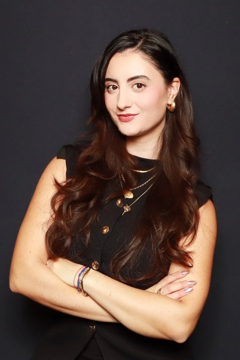 A woman with long dark brown hair, wearing a black sleeveless top, gold earrings, layered necklaces, and bracelets, standing with her arms crossed against a dark background.