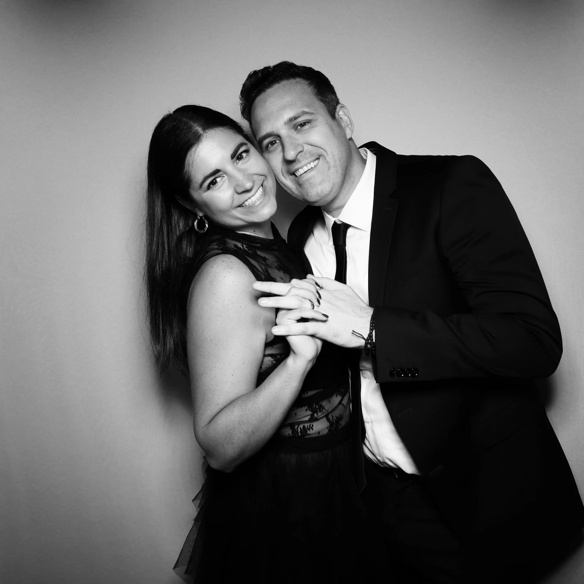 A happy couple in formal attire smiling and holding hands in front of a plain background.