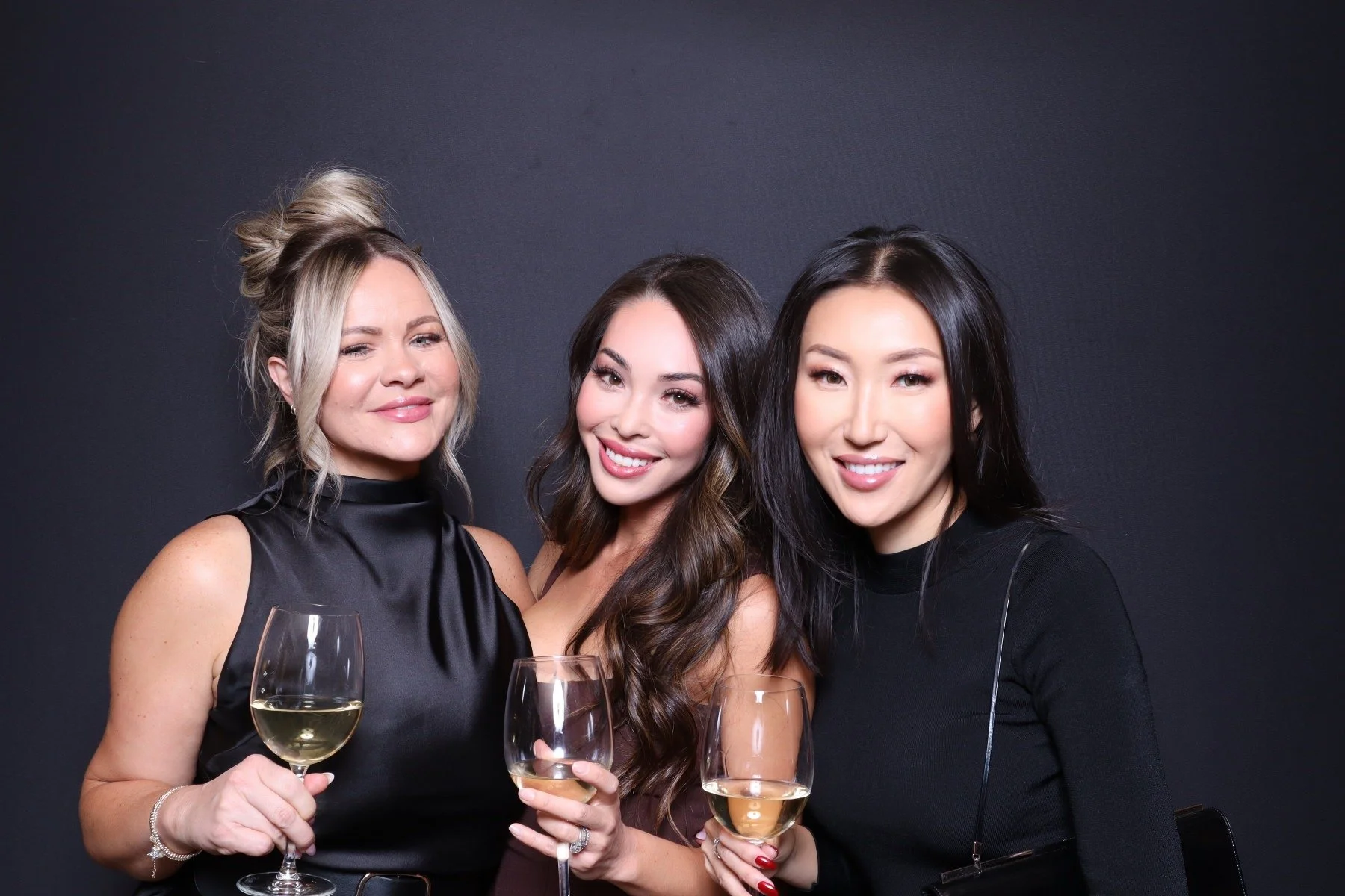 Three women smiling and holding glasses of white wine, standing against a dark background at a social gathering.