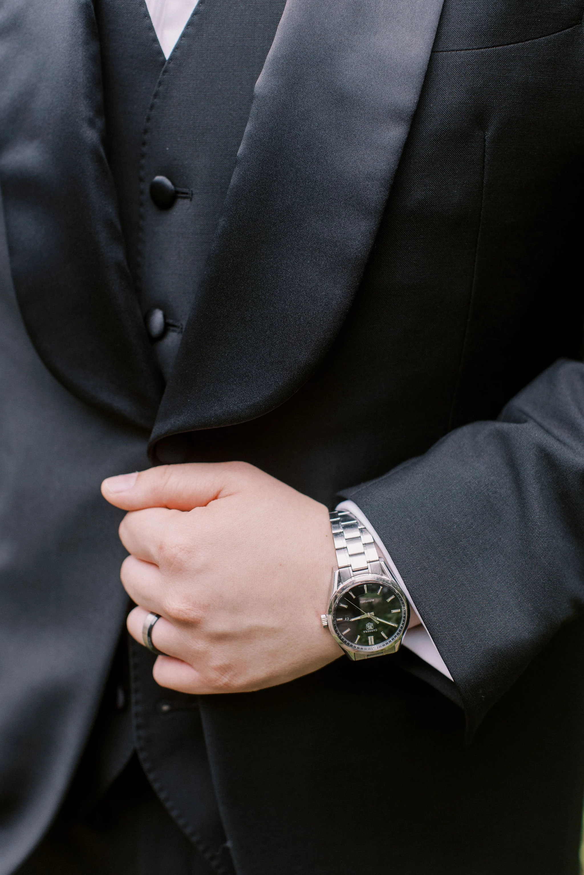 Close-up of a person wearing a black suit, with a silver watch on their wrist, and a wedding ring on their finger, holding their suit jacket.