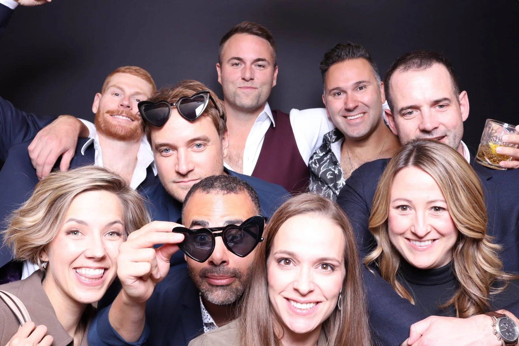 Group of ten people at a party, some holding drinks, posing against a dark background.