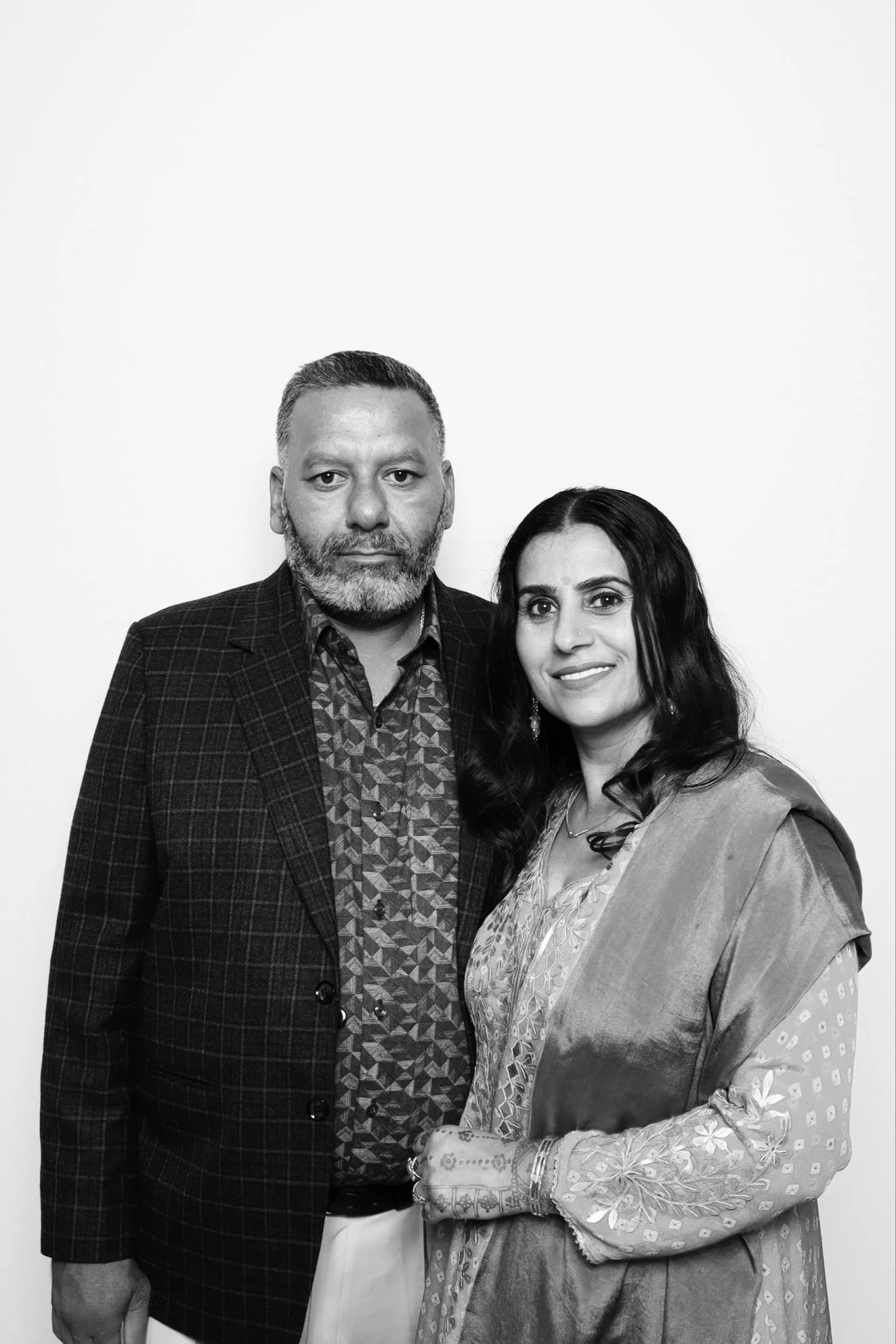 A black and white photograph of a man and woman standing close together, looking at the camera. The man has short hair and a beard, and is wearing a checkered blazer over a patterned shirt. The woman has long dark hair and is dressed in traditional I