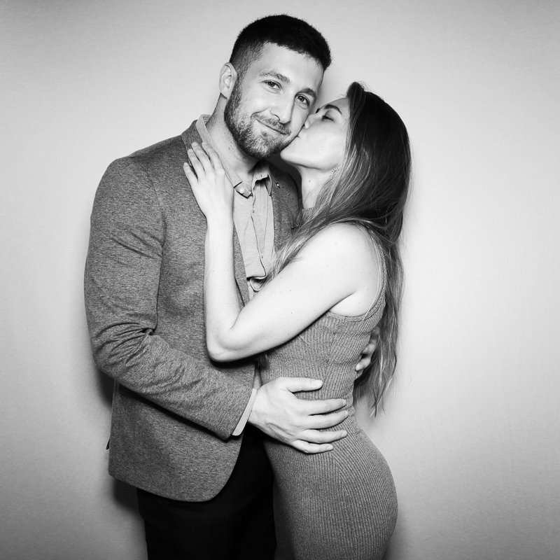 A black and white photo of a man and woman embracing. The woman is giving the man a kiss on the cheek, and he is smiling with a slight grin. The woman has long hair and is wearing a fitted sleeveless dress. The man has short hair, a beard, and is wea