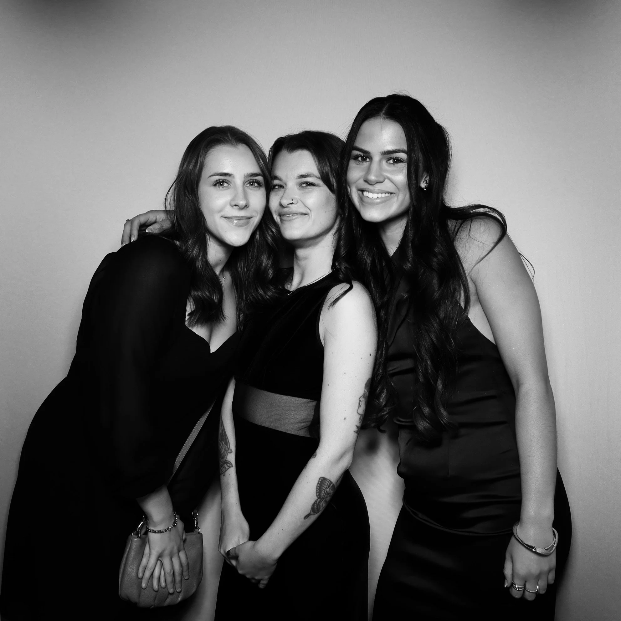 Three women dressed in black, smiling and standing close together against a plain background.