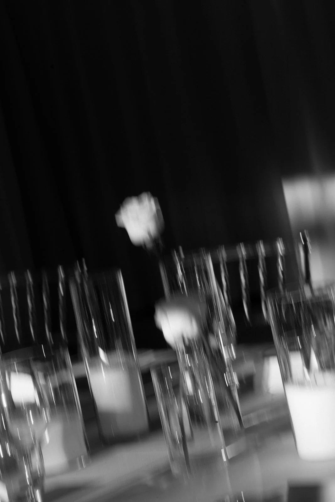 A black and white photo showing multiple empty glasses on a table, with a blurred background. There are two single white flowers in the scene.