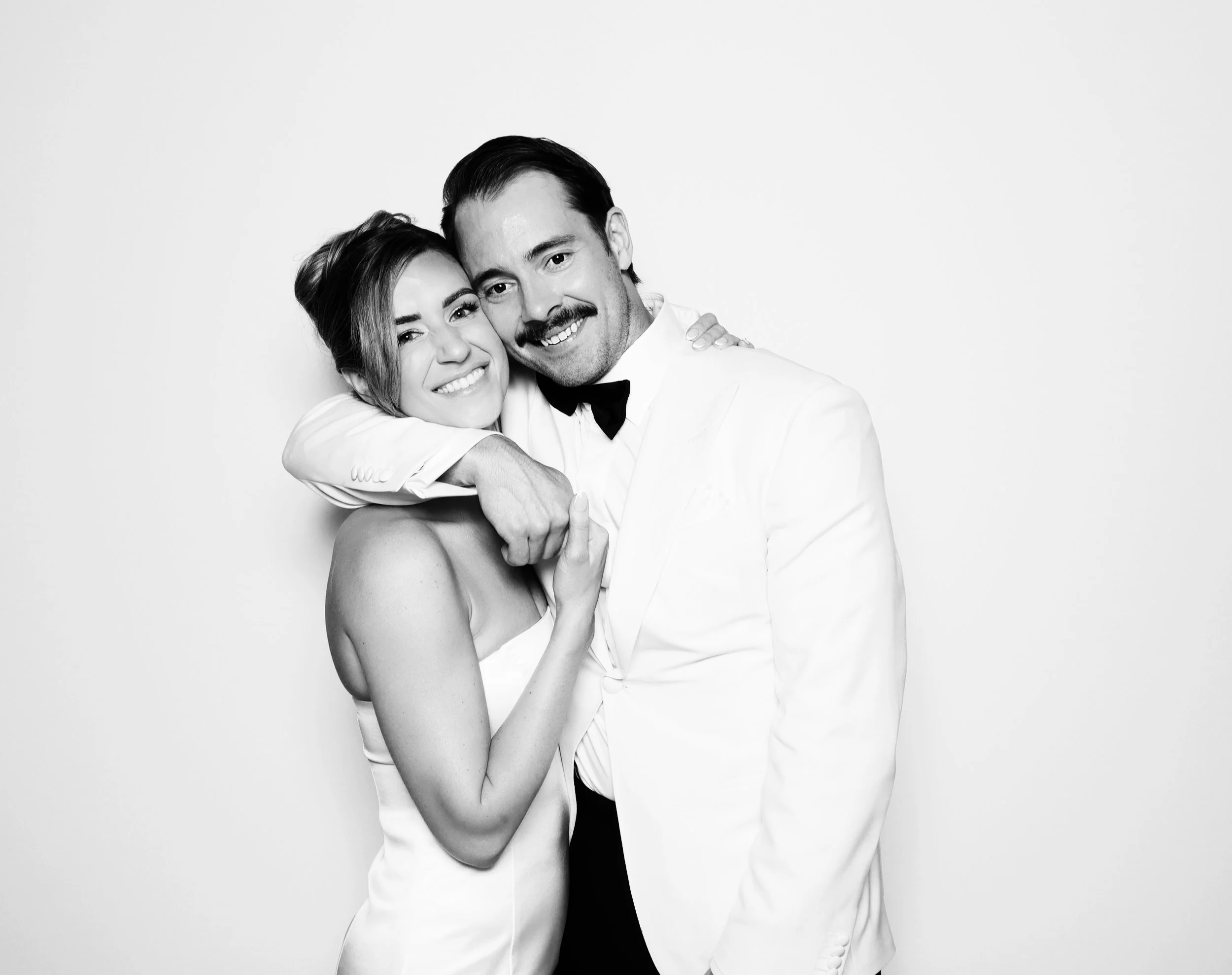 A black and white photo of a smiling couple dressed in formal attire, with the woman in a strapless dress and the man in a tuxedo, embracing each other happily.