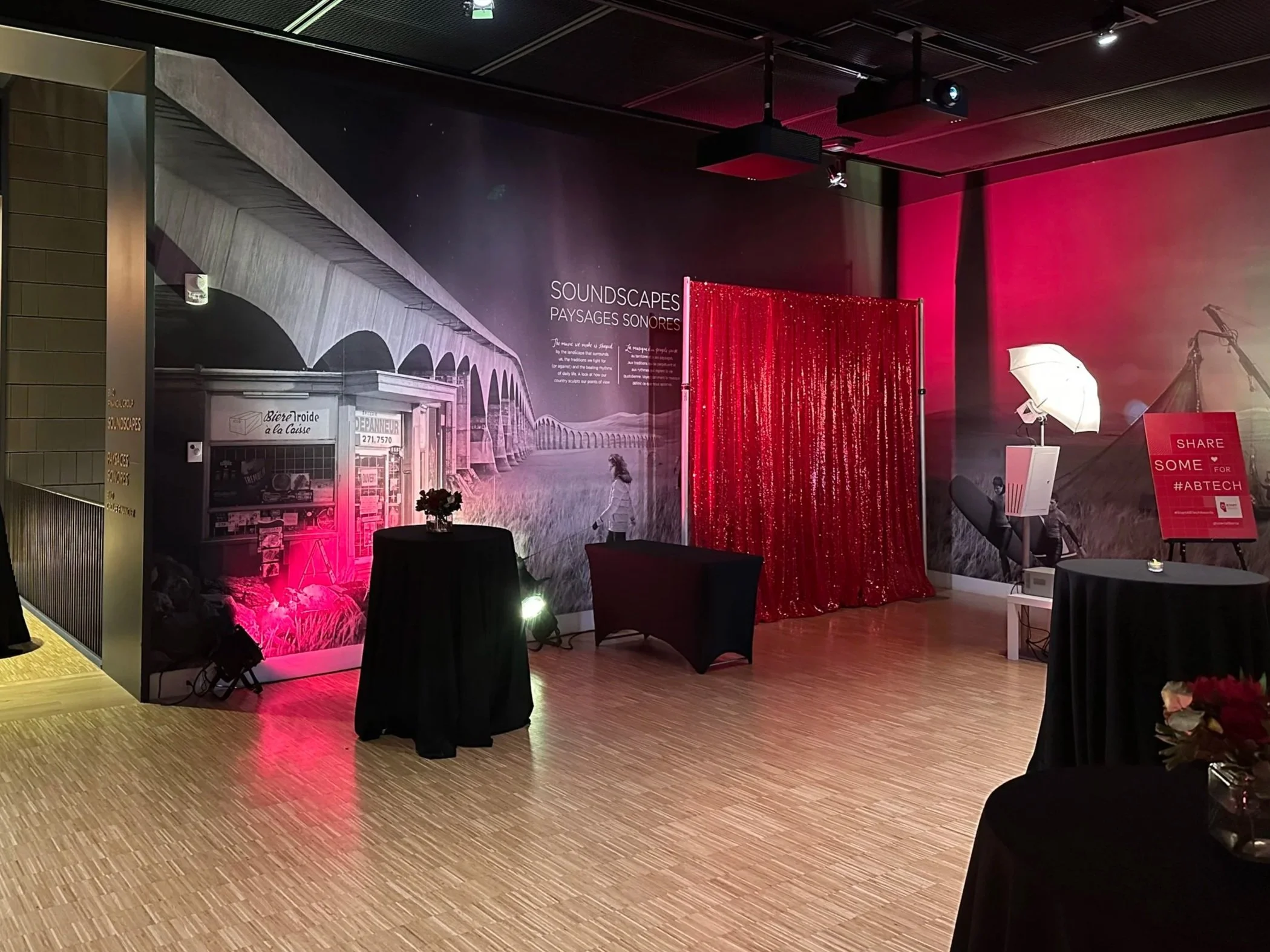 Event space with a mural wall titled 'Soundscapes Paysages Sonores', decorated with red and pink lighting and a red sequined curtain, and tables with black tablecloths, floral arrangements, and signs encouraging sharing for #ABTECH.