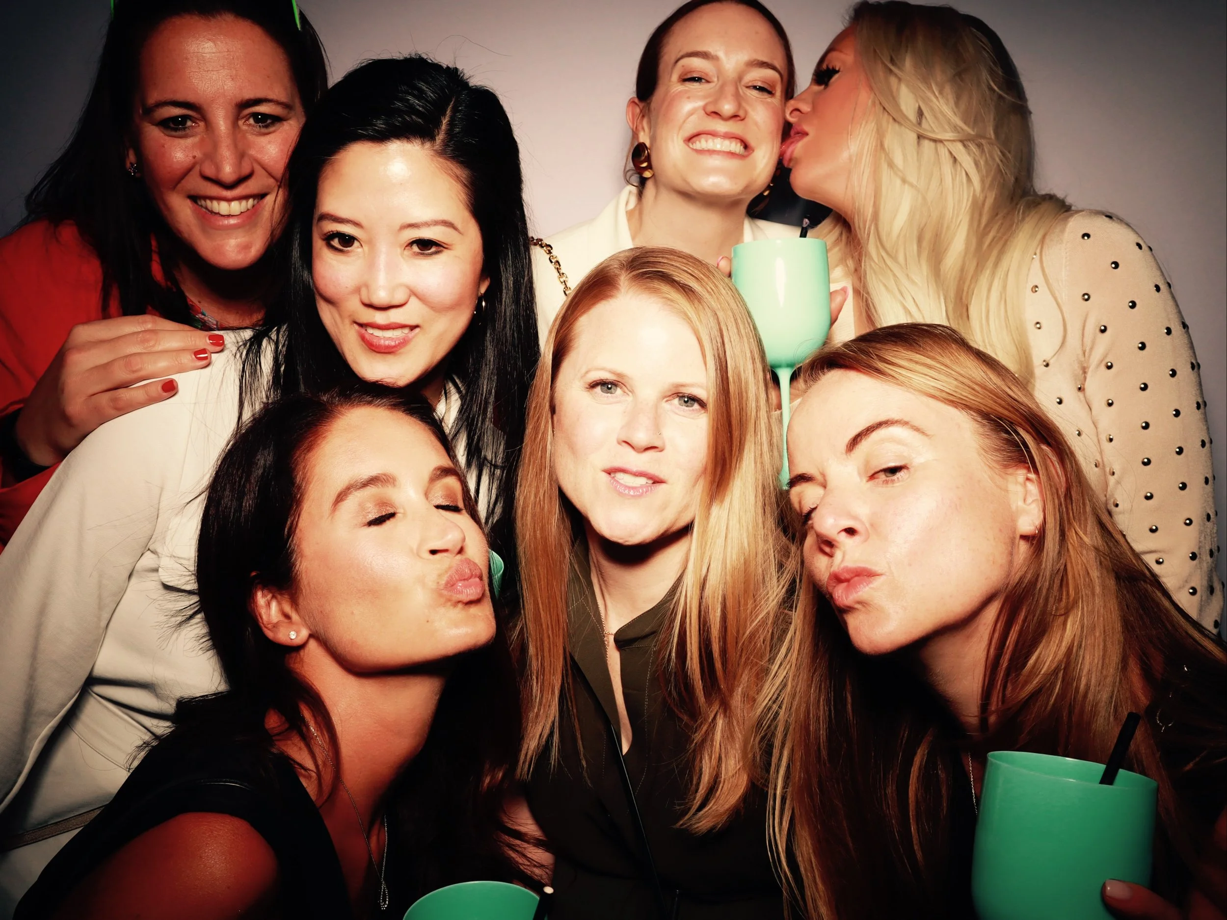Group of seven women at a party or gathering, some holding green cups, smiling, and making playful expressions.