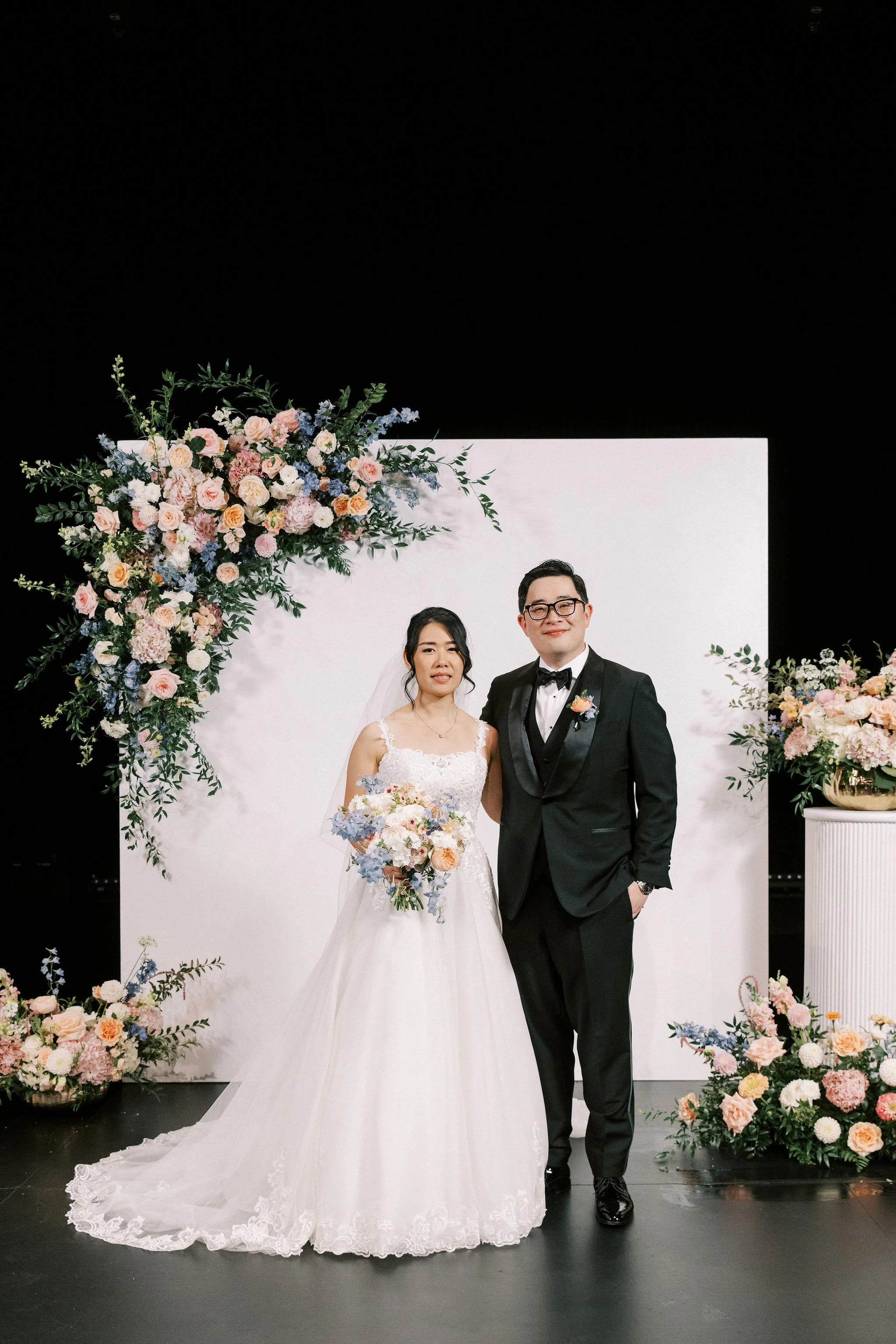 Bride and groom standing together at their wedding, with the bride holding a floral bouquet, standing in front of a white display with floral arrangements.