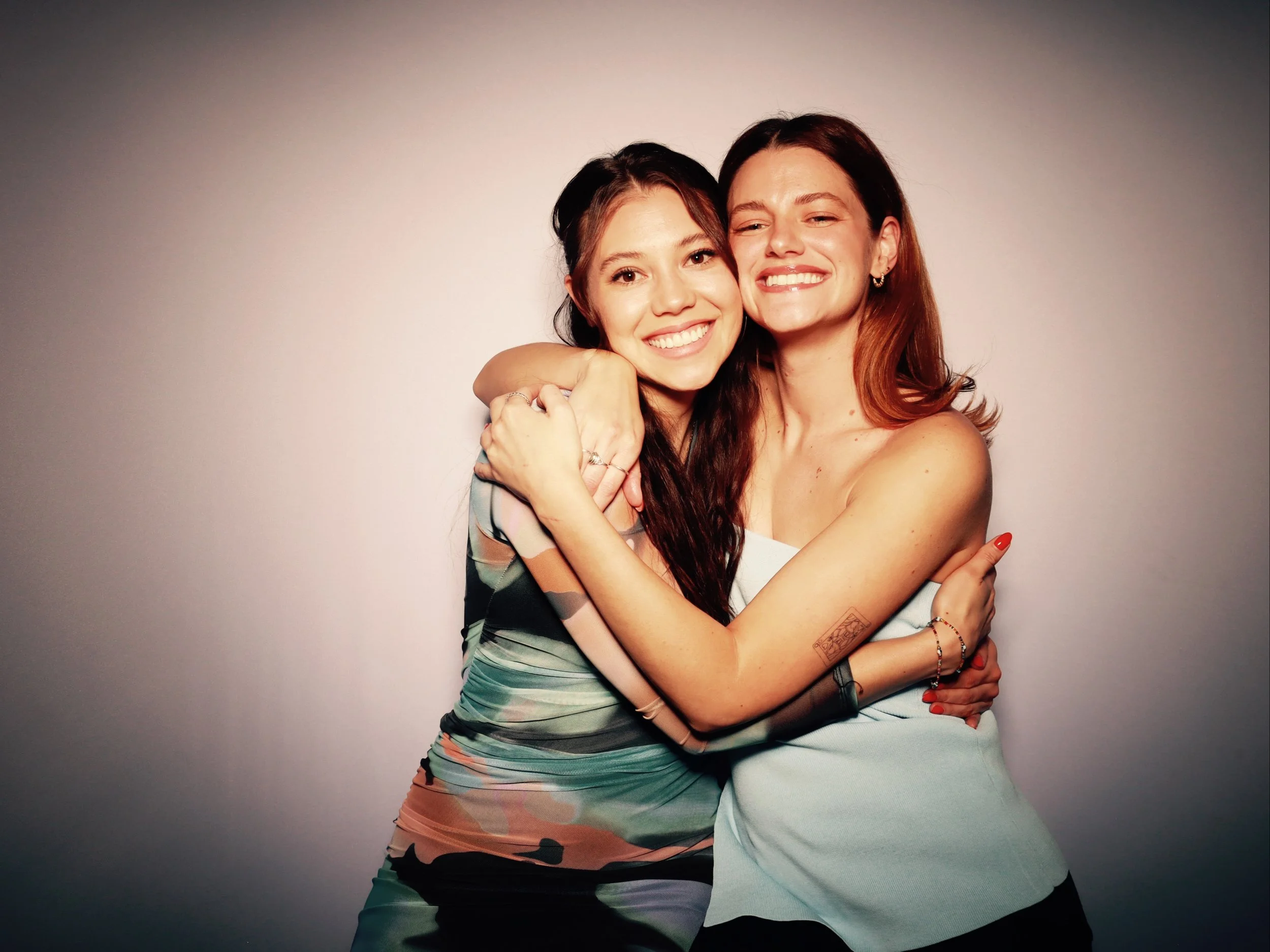 Two women hugging and smiling at the camera against a plain light background.