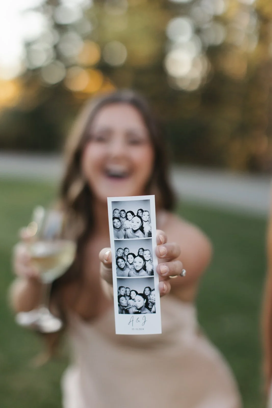 A woman dressed in white holding a four-photo photo booth strip in front of her, with her face blurred in the background. She is outdoors with trees and bokeh lights, holding a glass of champagne in her other hand.