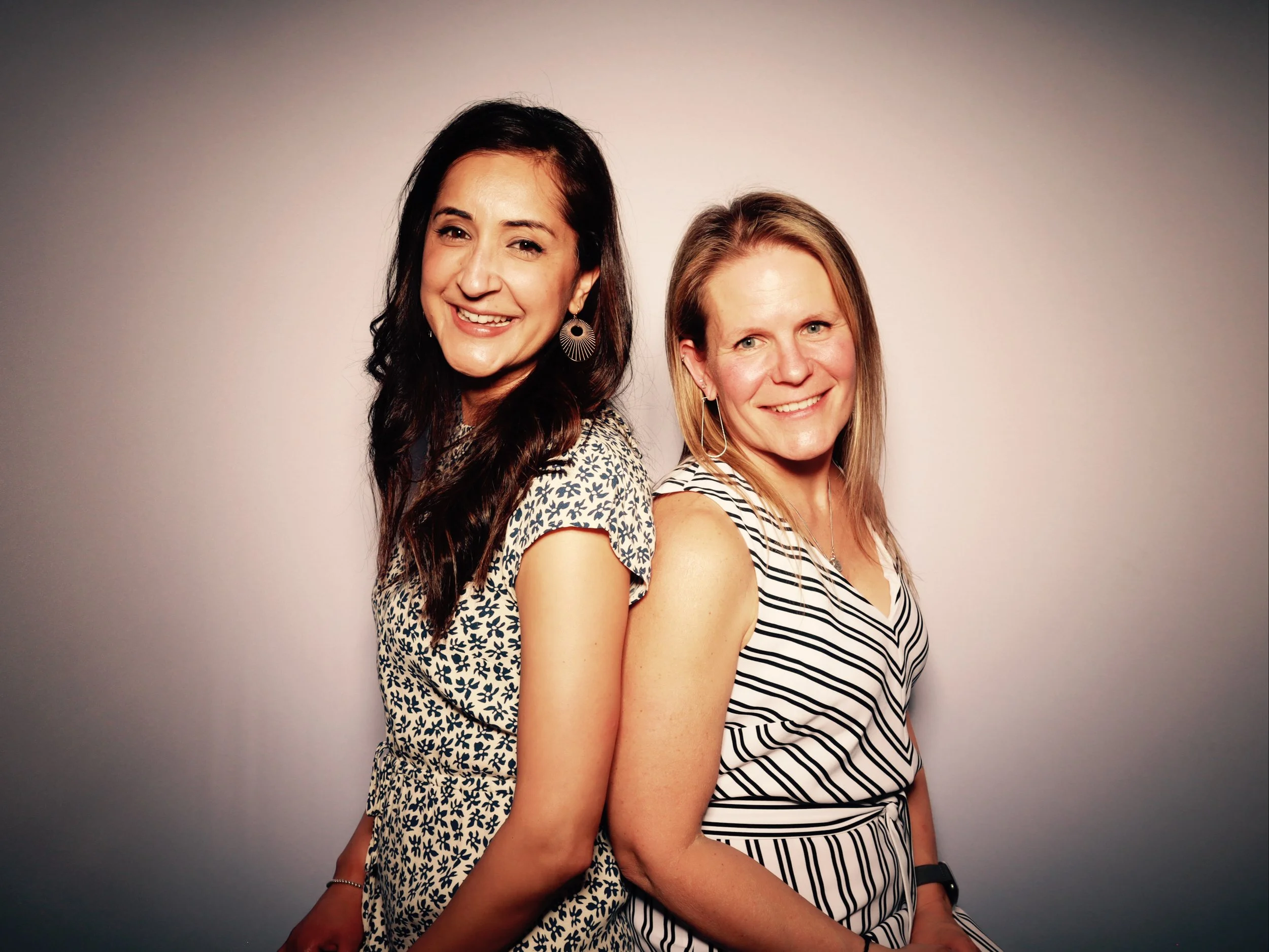 Two women standing back-to-back, smiling, in front of a plain gray background.