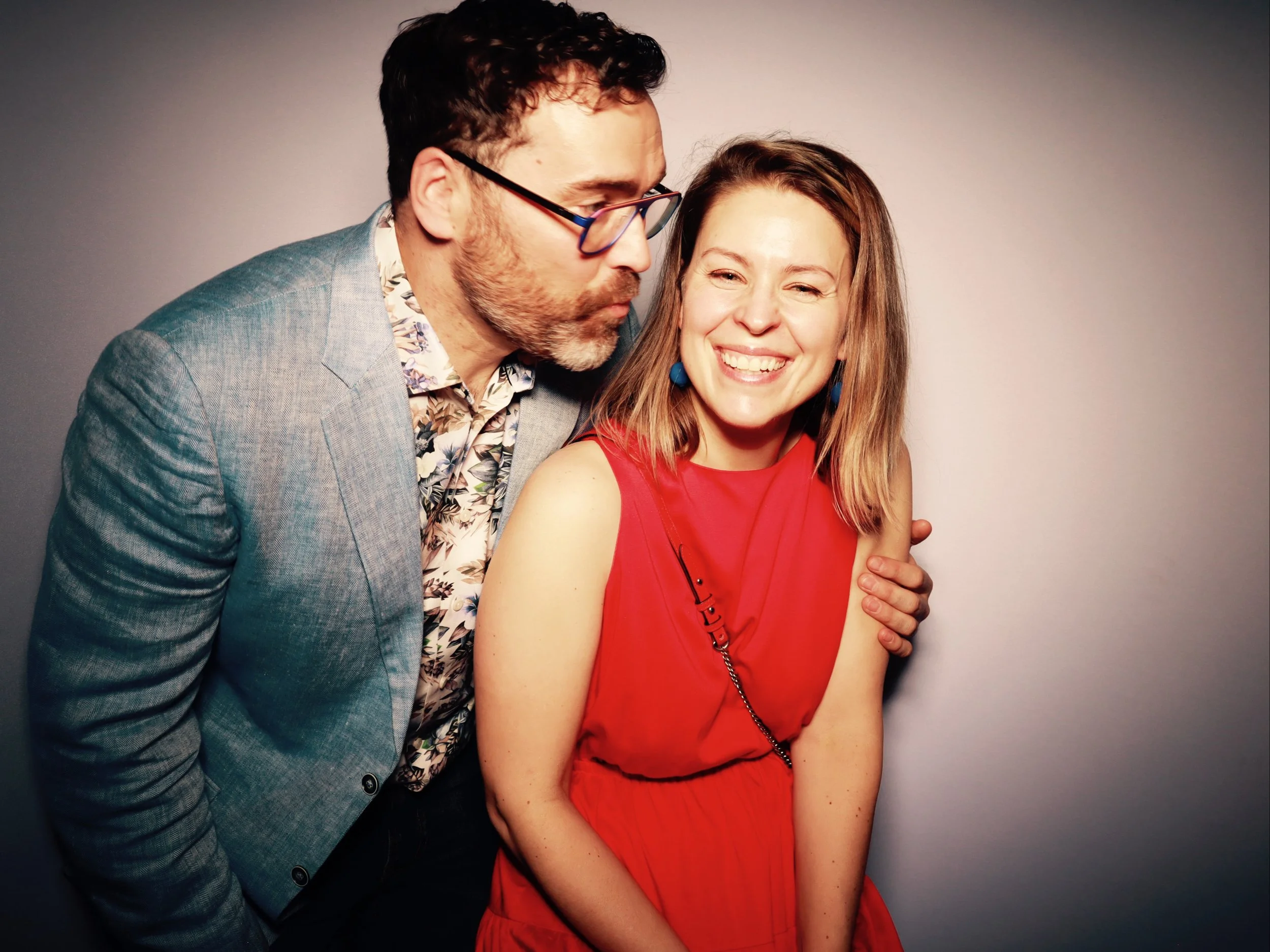 A man in glasses and a light gray blazer leans in to kiss a woman in a red dress, who is smiling.