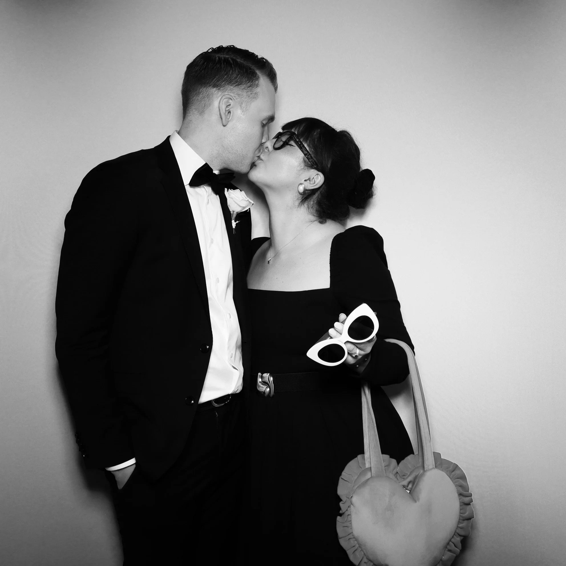 A man and woman in formal attire kissing, with the man in a tuxedo and the woman holding a pair of white sunglasses and a heart-shaped handbag.