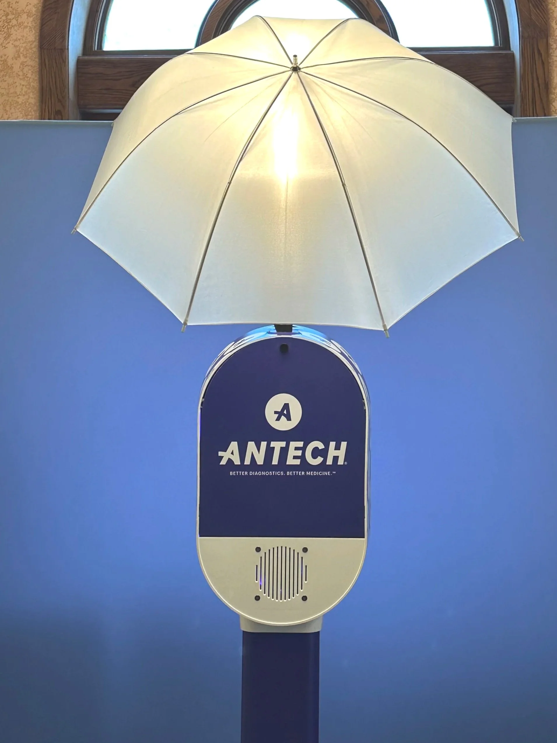 A hand sanitizer dispenser with the logo and name 'Antech' printed on it, mounted on a stand, with a yellow umbrella placed above it for coverage, located indoors near a window.