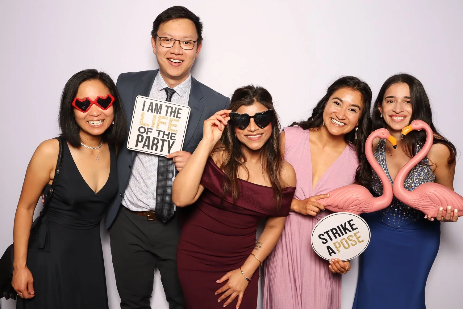 A group of five diverse people dressed up and smiling at a photo booth, holding signs and props such as heart-shaped sunglasses, flamingo inflatables, and signs that read "I am the life of the party" and "Strike a pose."