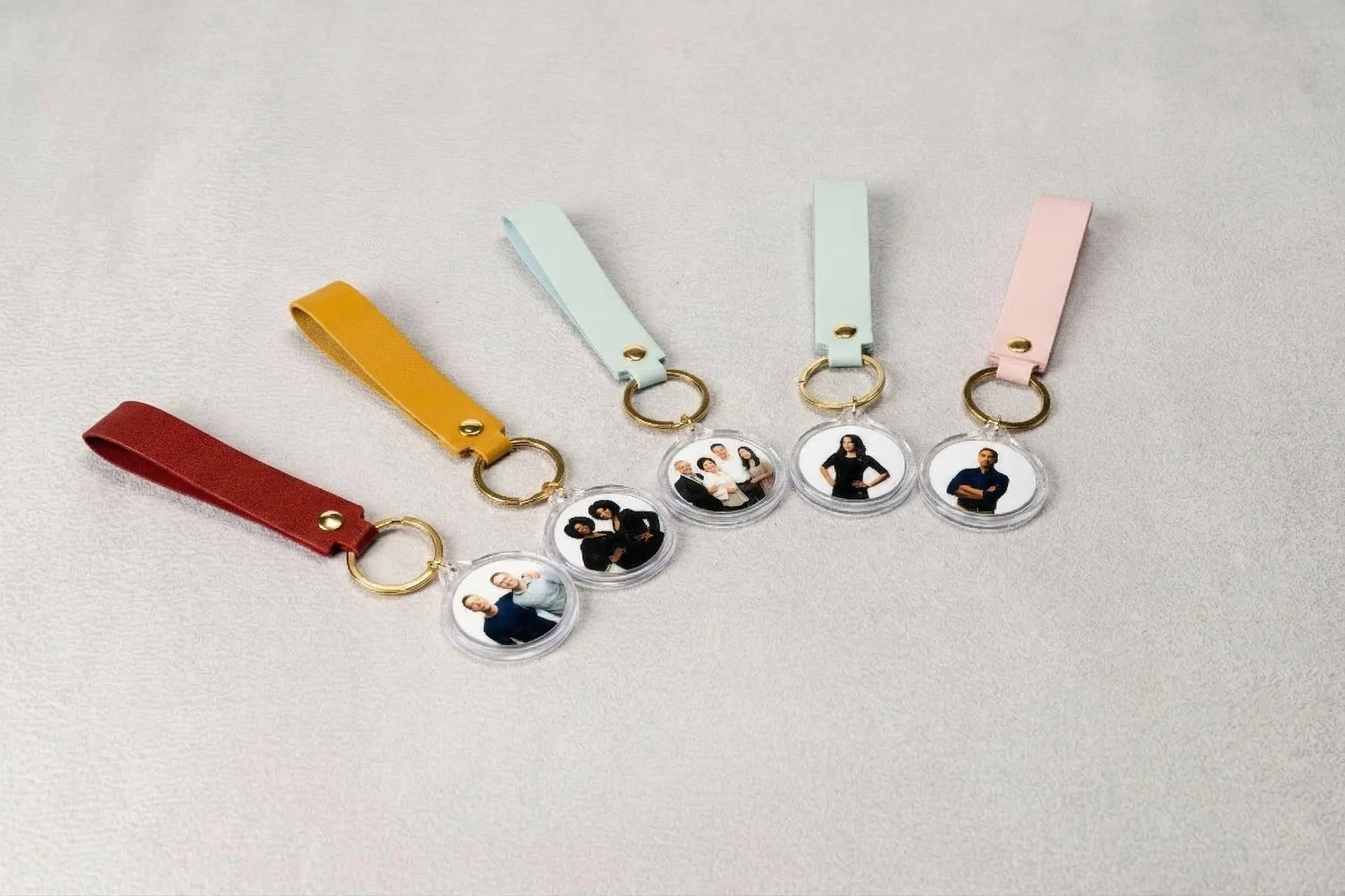 Five keychains with colorful leather straps and circular photo holders, each displaying a different group or individual portrait, arranged on a light surface.