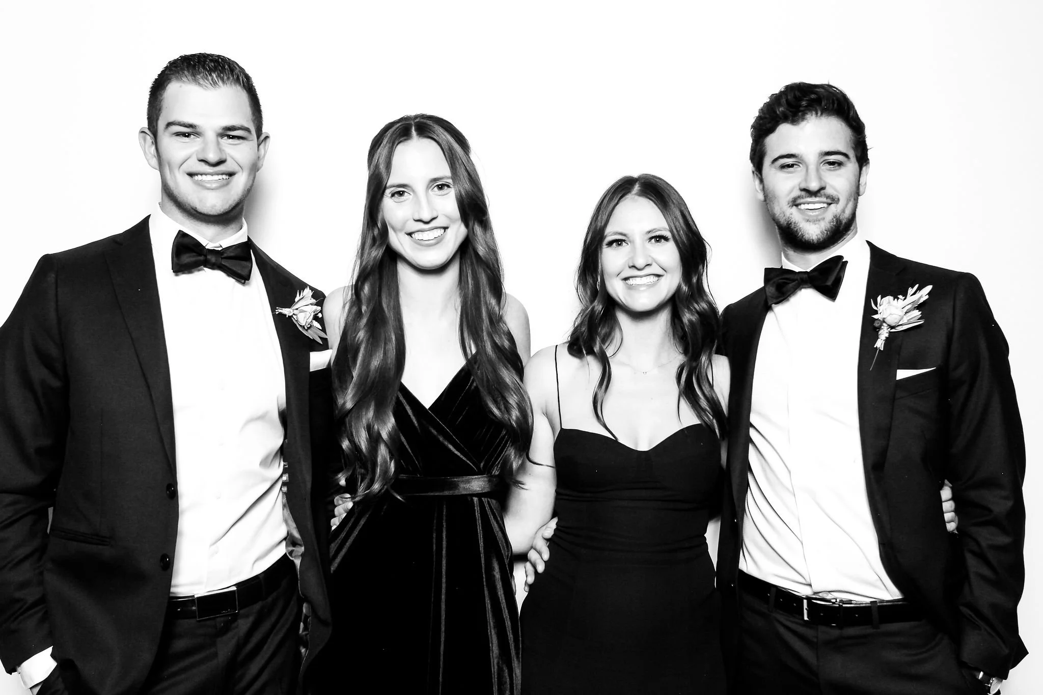 Four young adults in formal attire posing together against a plain white background, smiling at the camera. The men are wearing tuxedos with bow ties and boutonnières; the women are dressed in elegant dark dresses.