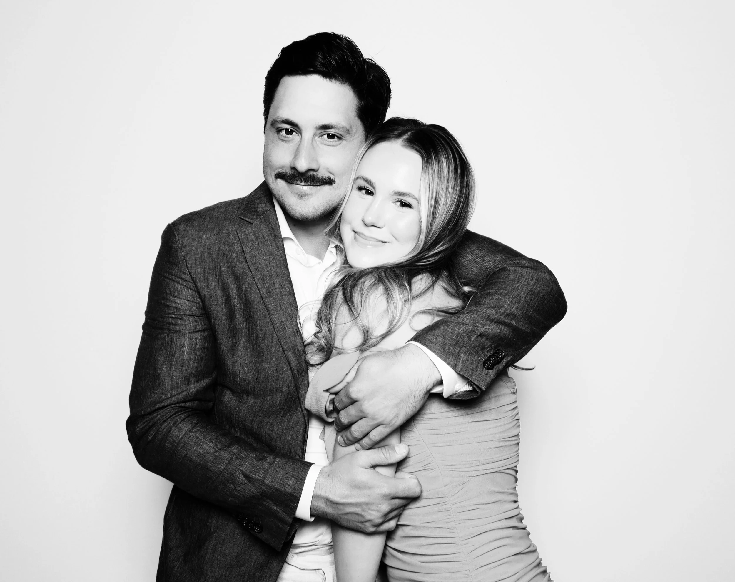 Black-and-white photo of a smiling man and woman hugging. The man has dark hair, a mustache, and is wearing a suit. The woman has long wavy hair and is wearing a sleeveless dress.