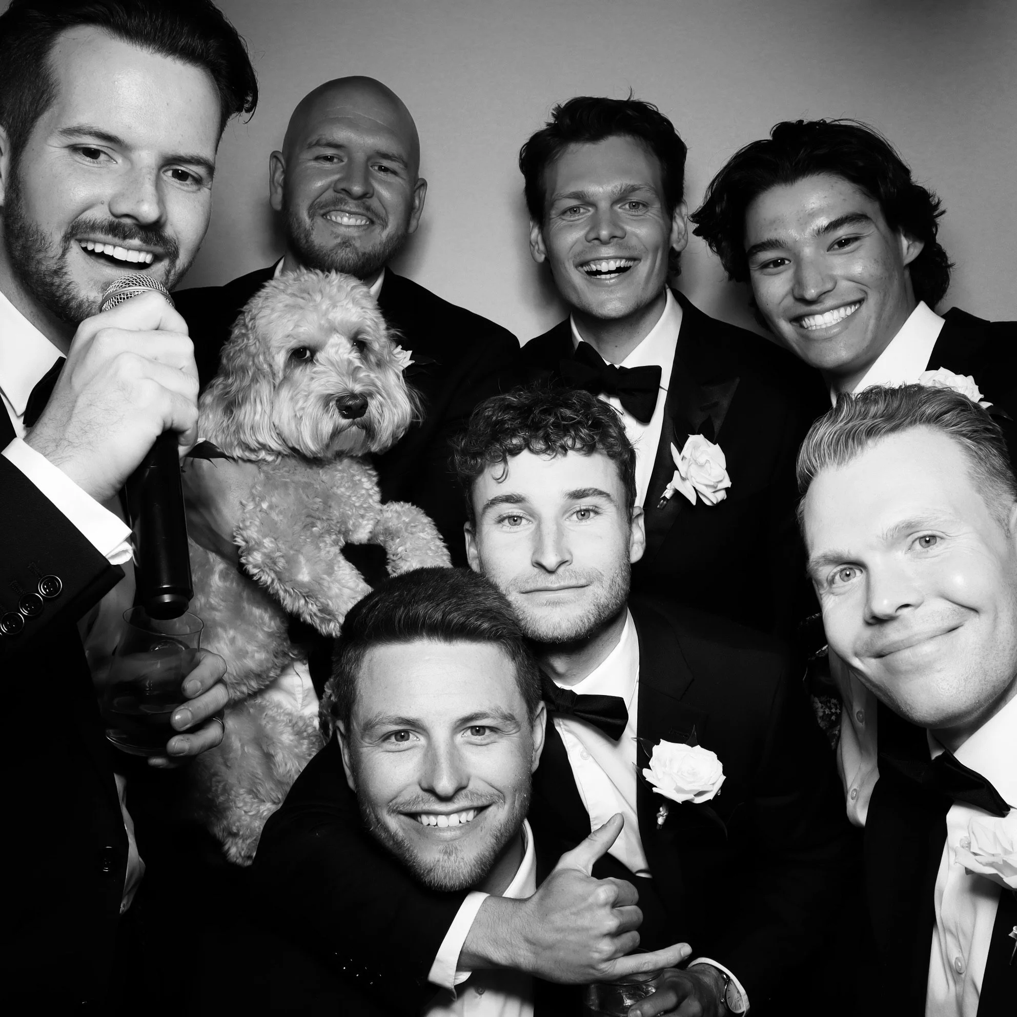 A group of seven men in tuxedos and bow ties, with one man holding a small, curly-haired dog, all smiling and posing together at a formal event, black and white photograph.