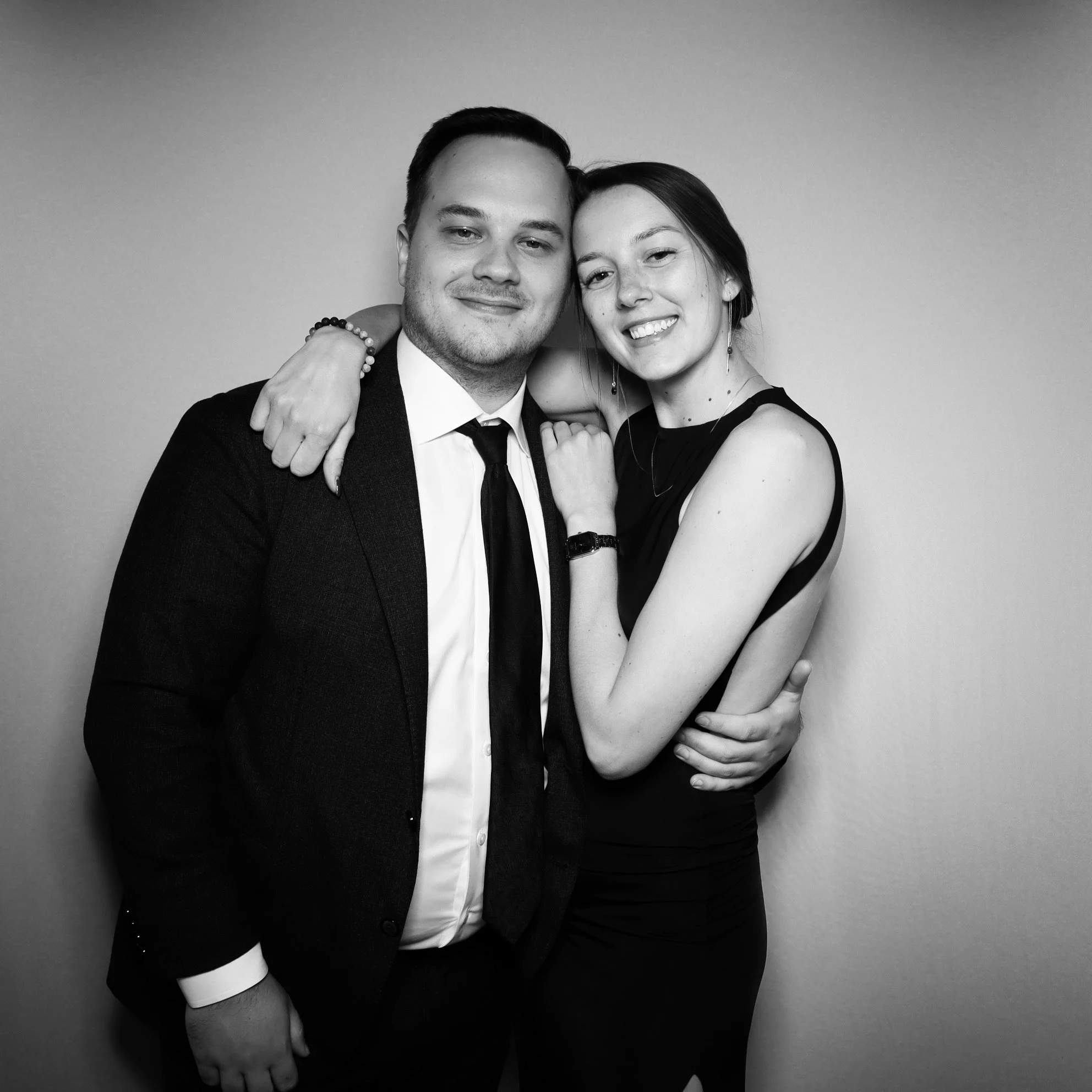 A black and white photo of a smiling man and woman embracing each other, dressed formally, against a plain background.