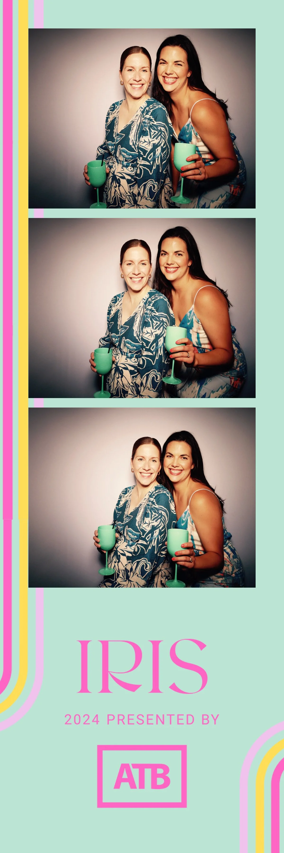 Three women smiling and posing together in a photo booth, each holding a turquoise wine glass. They are dressed in summery outfits and standing against a plain gray background.