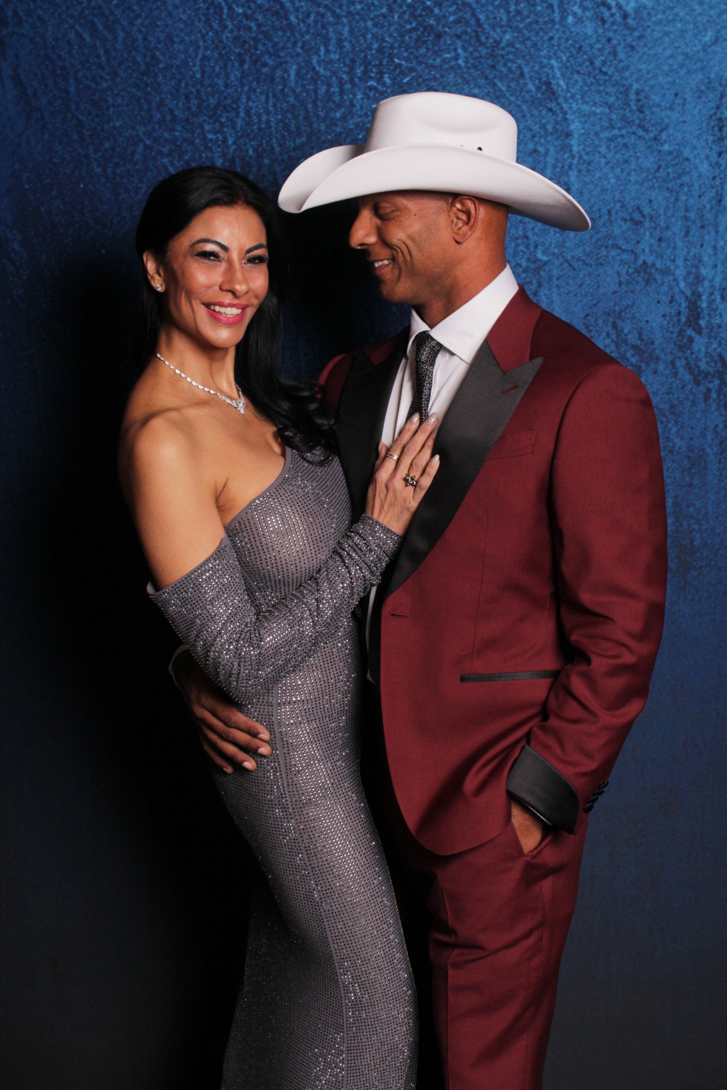 A smiling woman in a sparkly, off-shoulder, gray evening gown and a man in a burgundy tuxedo with a white cowboy hat, standing against a blue background.