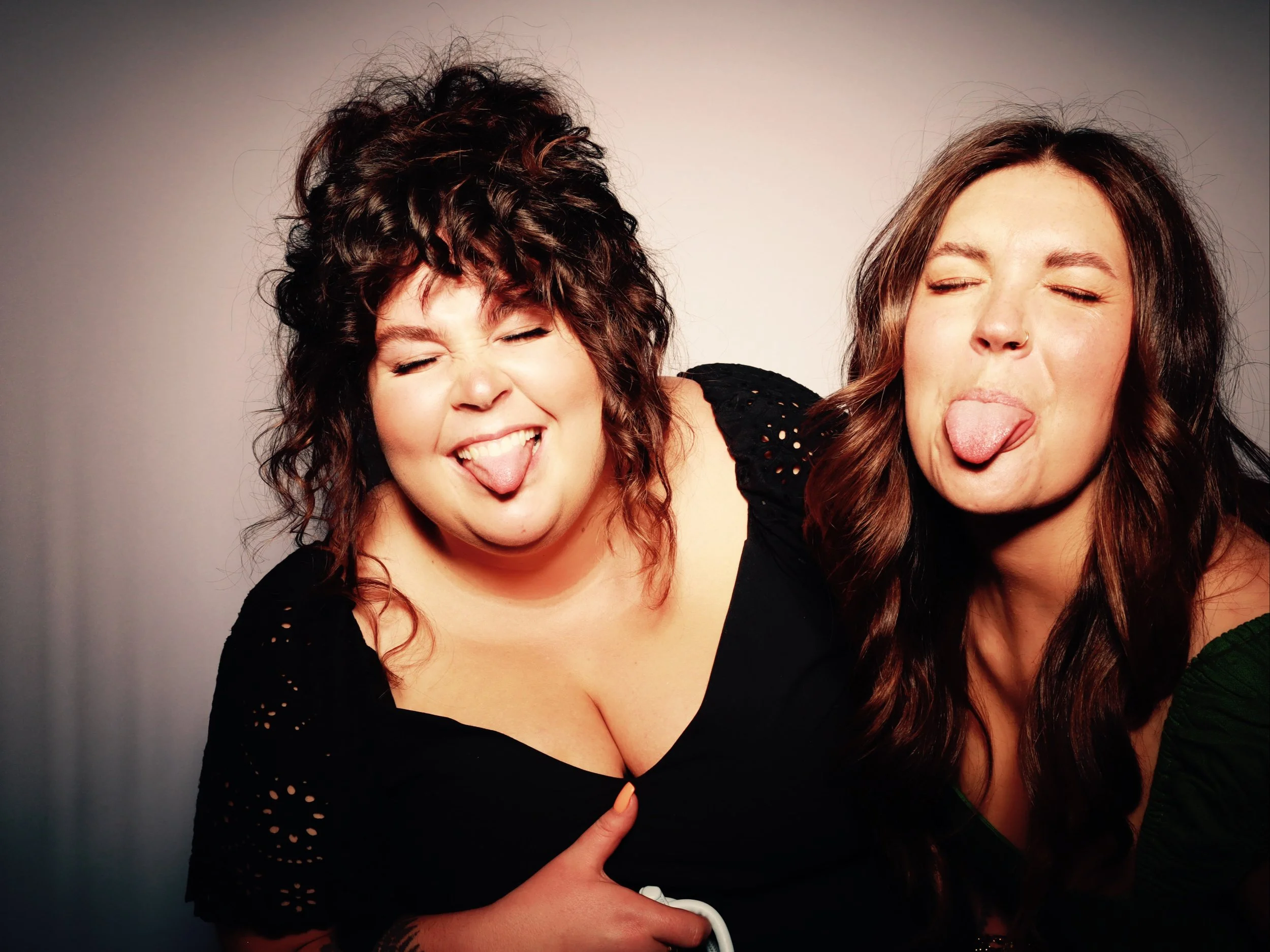 Two women with long hair making silly faces, sticking out their tongues and closing their eyes, against a plain background.