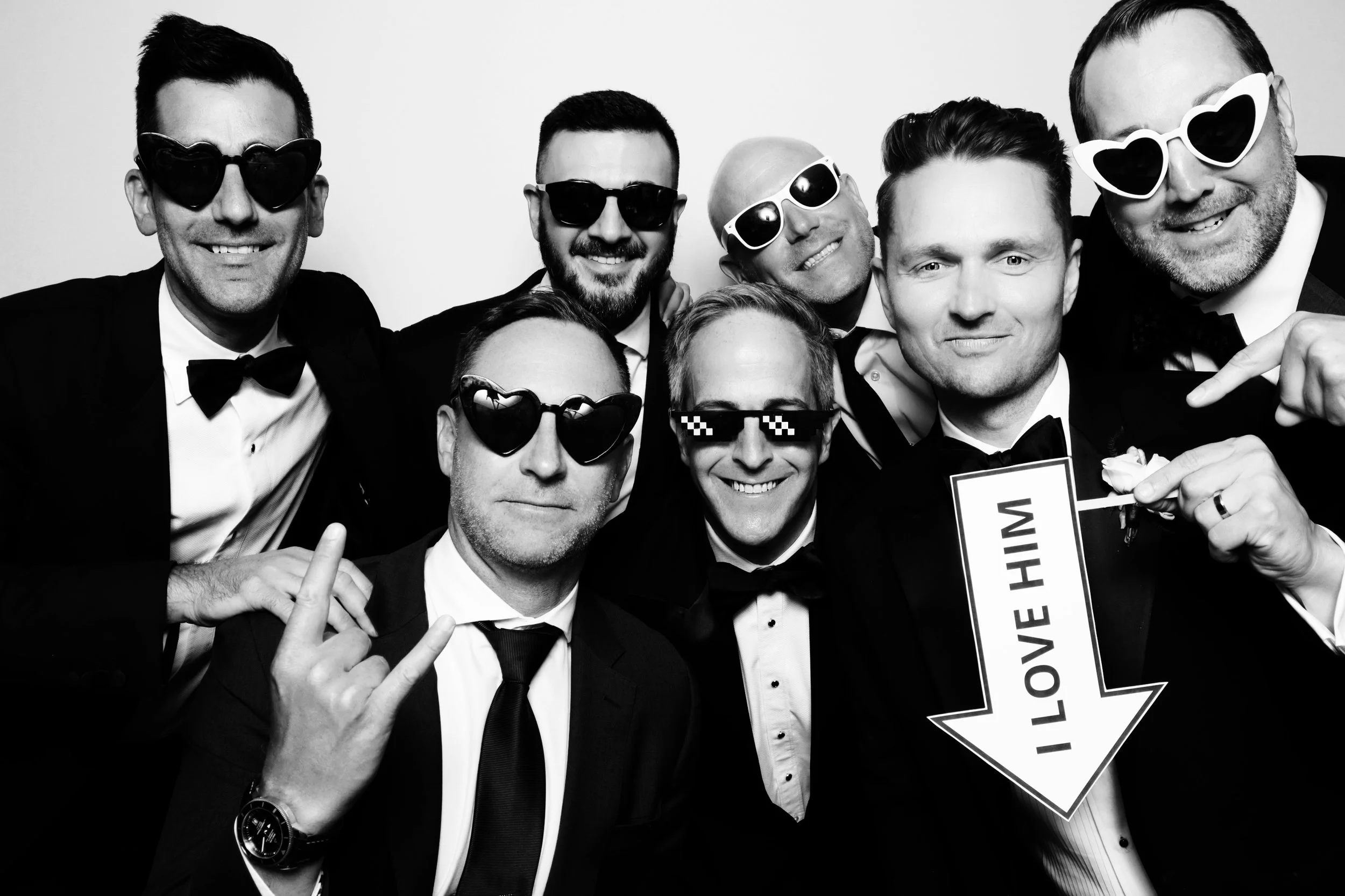 Group of men in tuxedos and sunglasses posing for a photo with one holding a sign that says 'I love him' pointing downward.