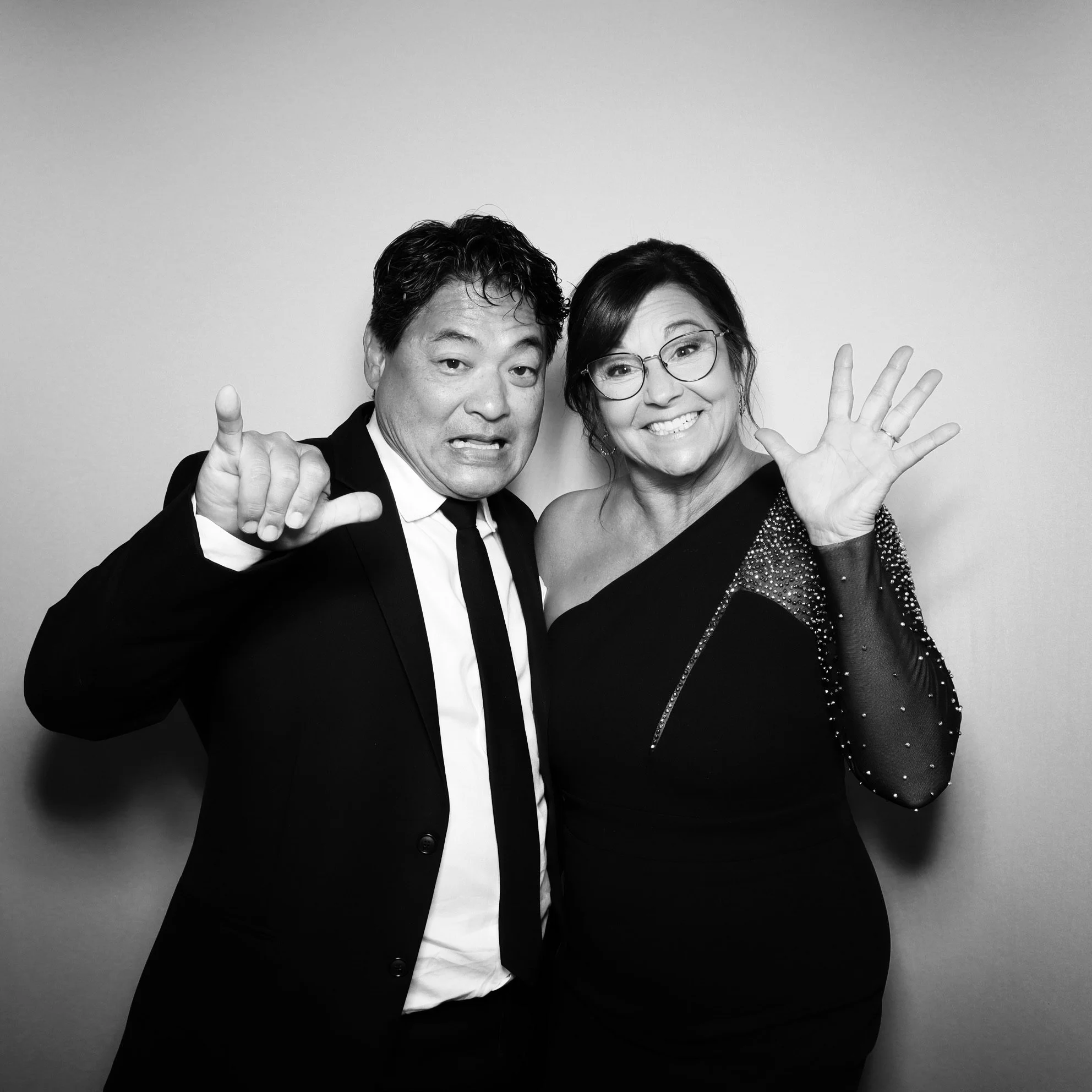 A man and a woman posing together, both smiling and making playful gestures, in black and white.