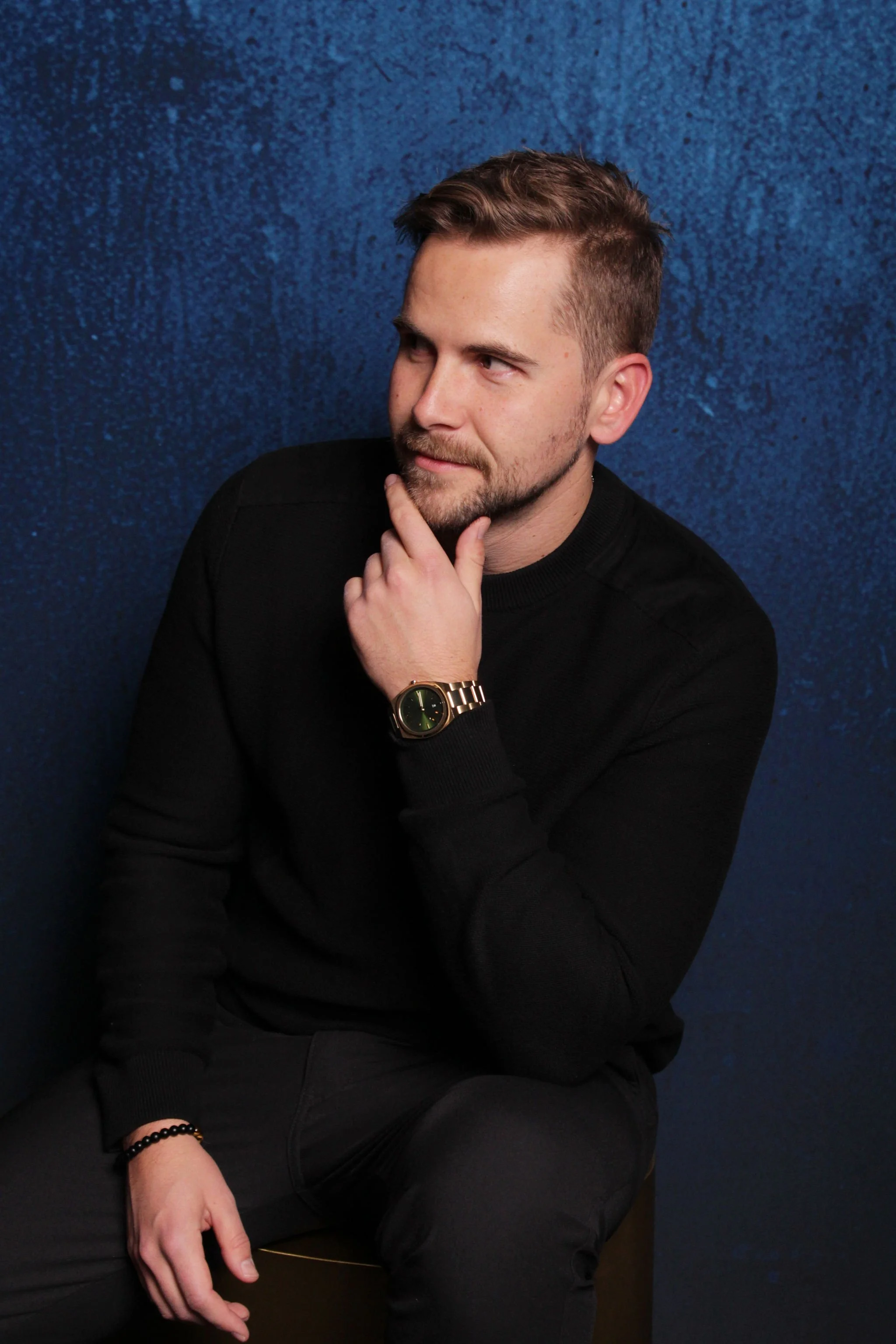 A young man with short brown hair, a beard, and wearing a black long-sleeve shirt, sitting against a textured blue background, has a thoughtful pose with his hand on his chin and a slight smile.